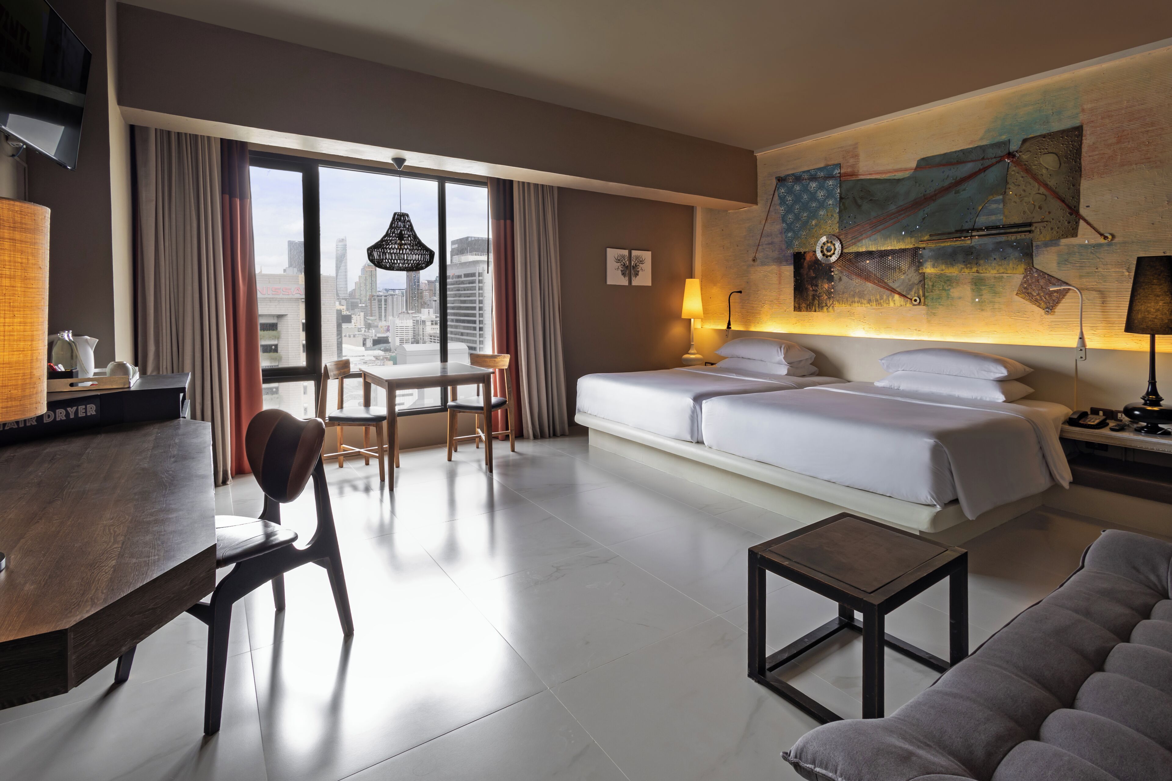 A hotel room with a bed, a desk, a chair, a small table, a sofa, a large window with curtains, and a city view.