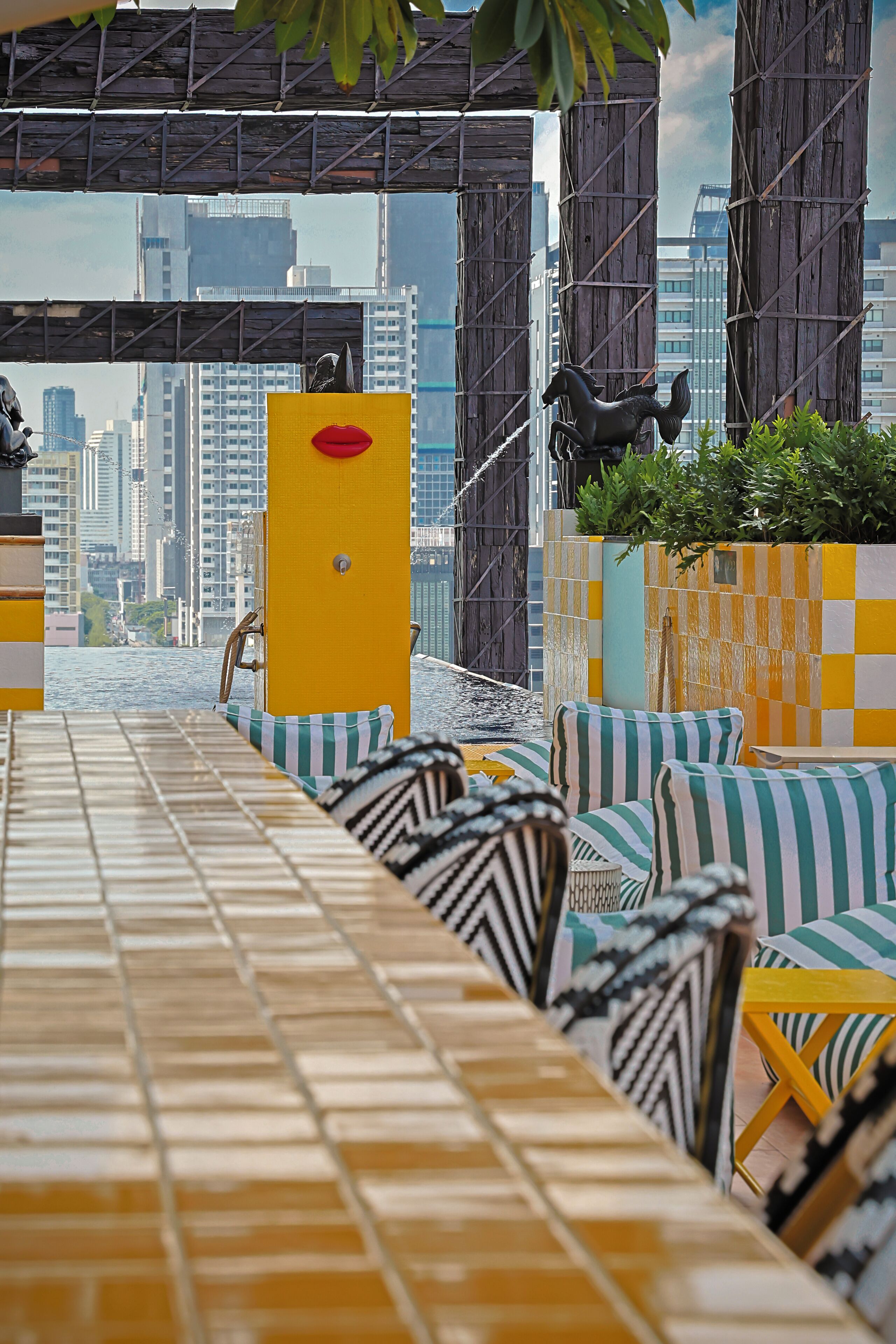 Un bar en la azotea con vistas a la ciudad, asientos a rayas y un pilar amarillo.