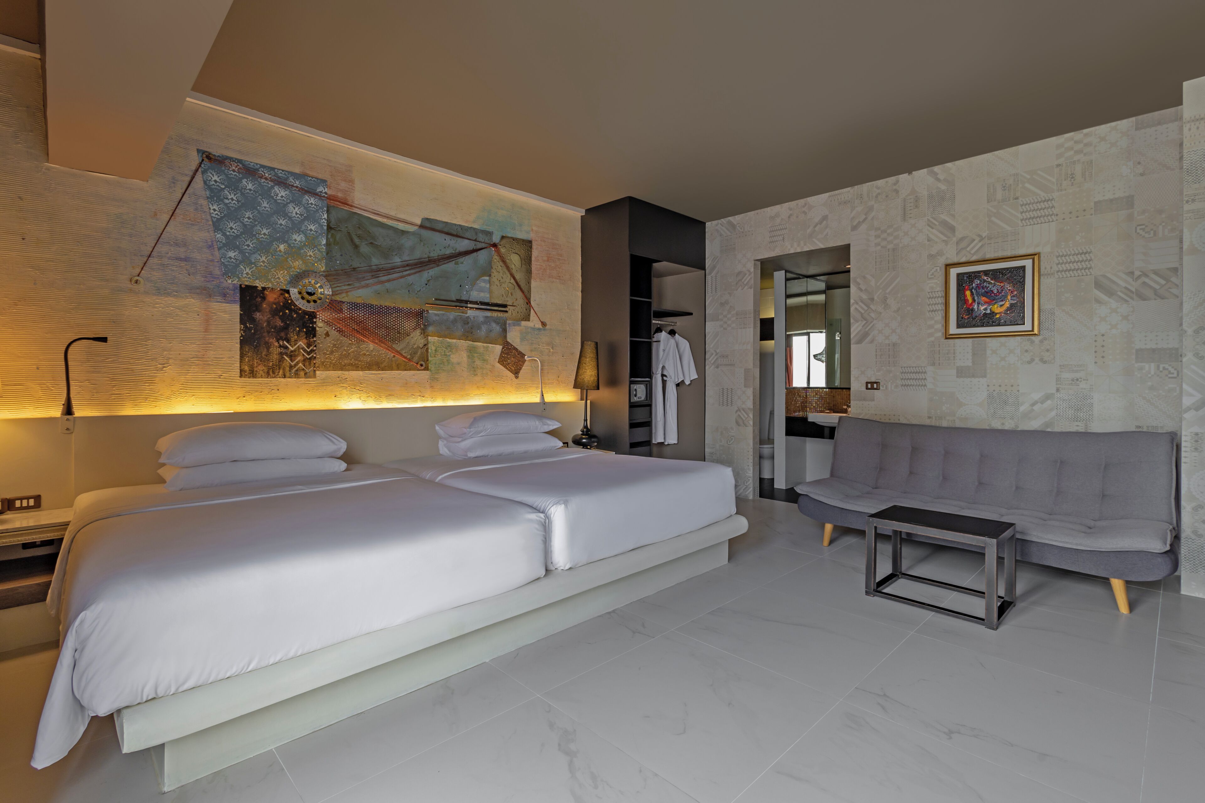 A modern hotel room with a large bed, a sofa, a small table, and a wall with abstract art.