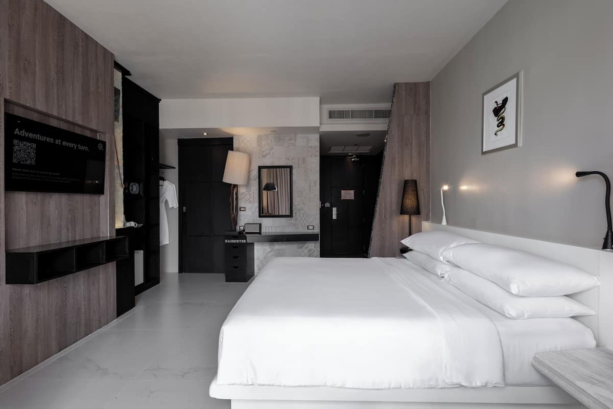 A modern hotel room with a large bed, a vanity with a mirror, a wall-mounted TV, and a framed artwork.