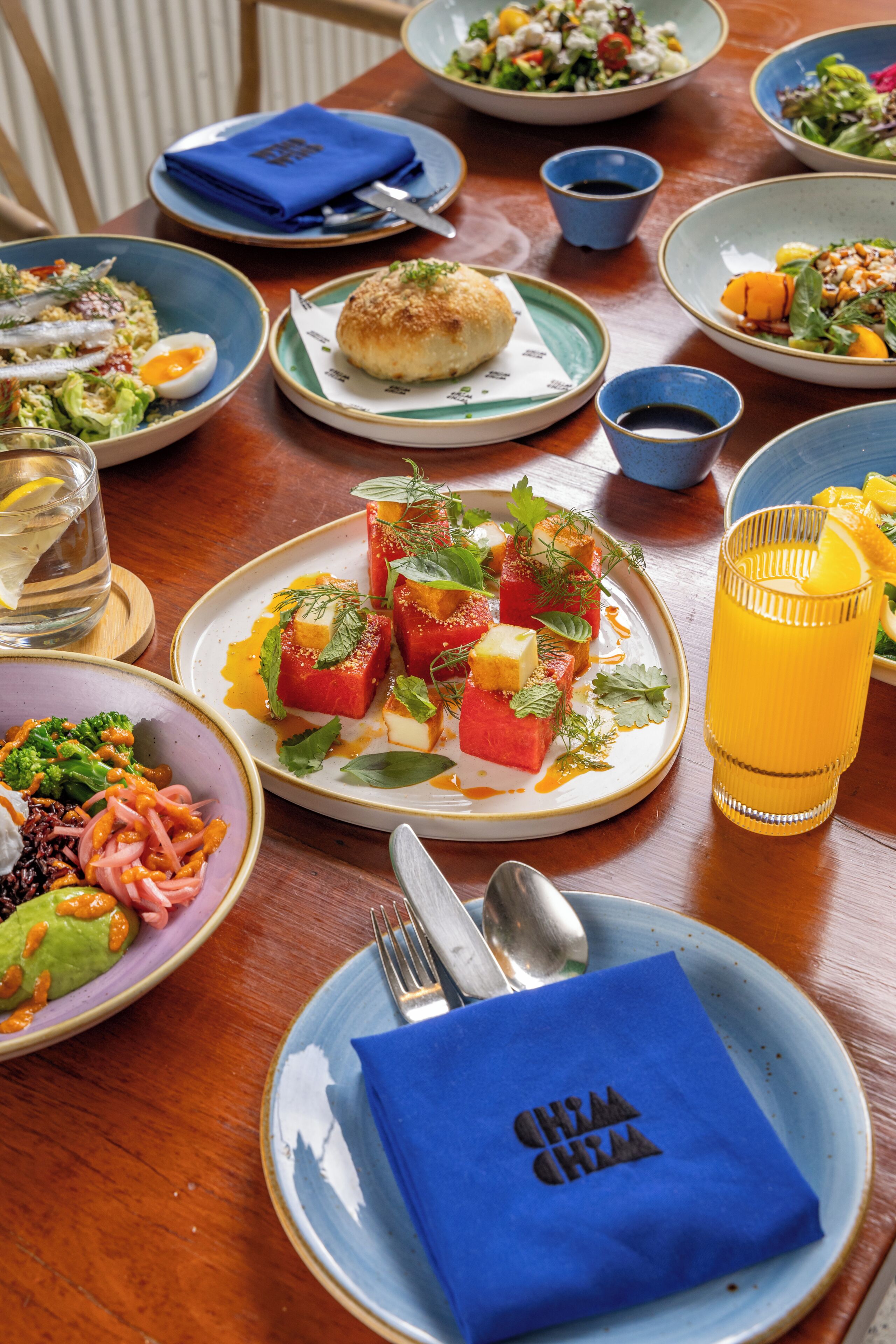 A dining table with various dishes, a glass of orange juice, and a blue napkin with a logo.