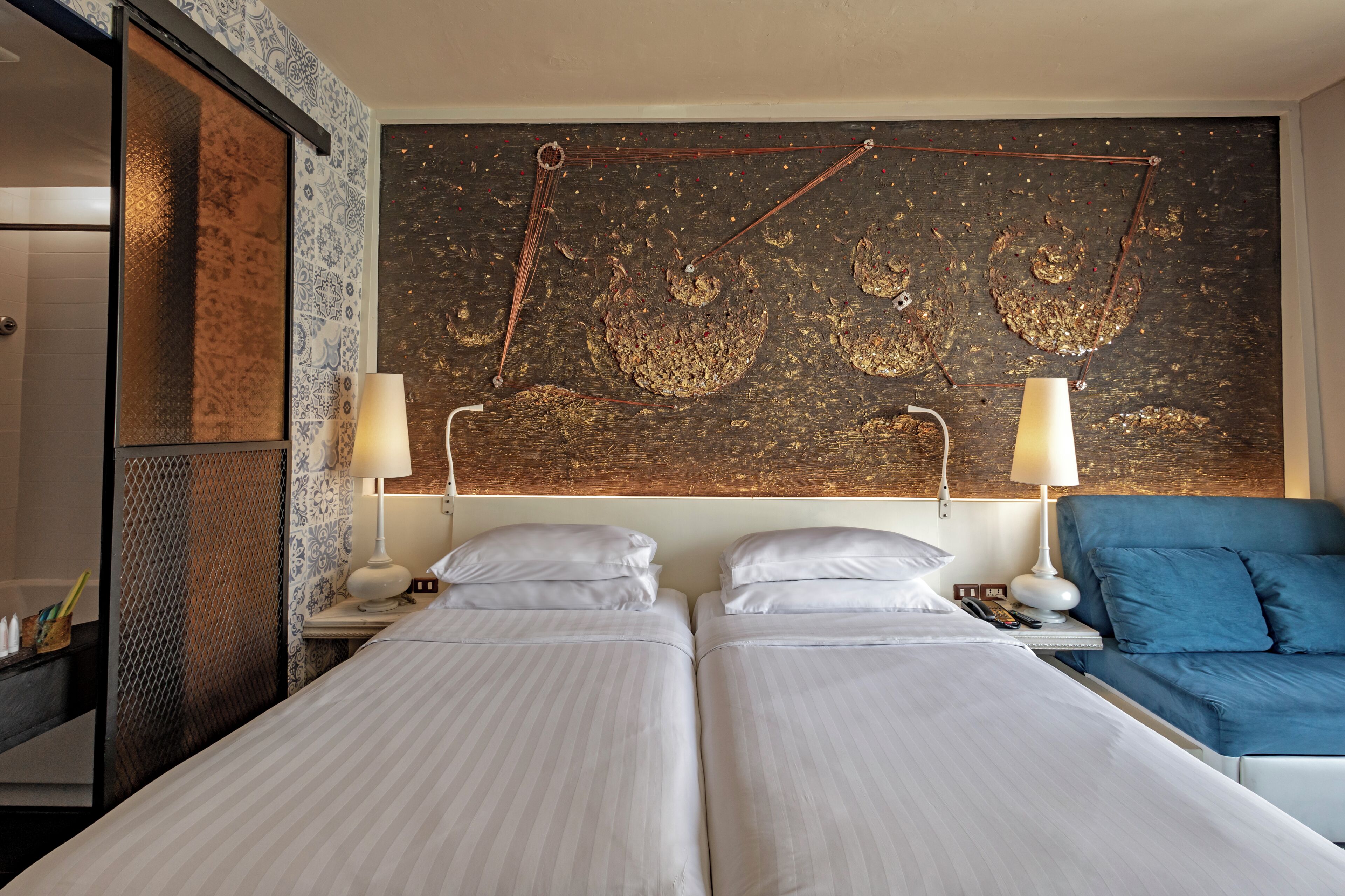 A hotel room with two beds, a large abstract artwork on the wall, and a blue armchair.
