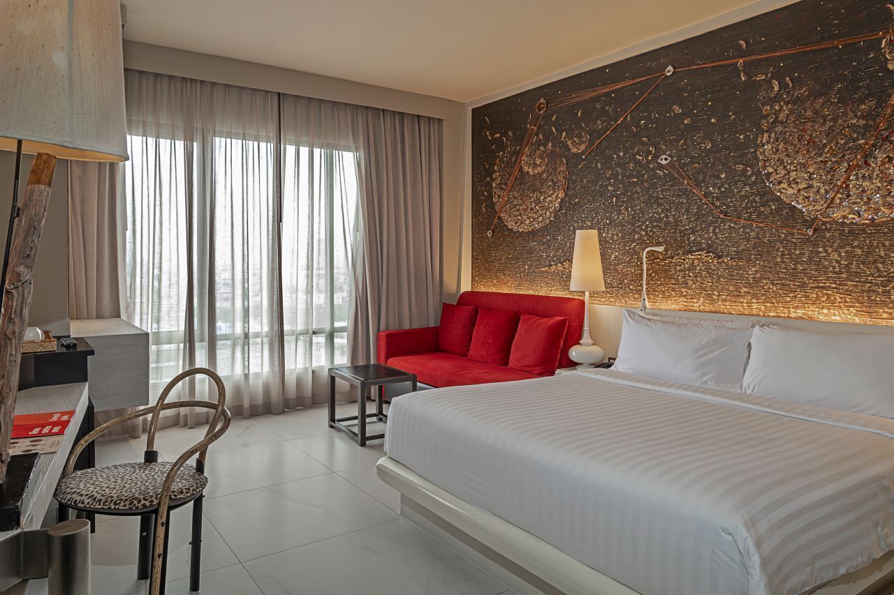 A hotel room with a bed, a red sofa, a desk, a chair, and a large abstract painting on the wall.