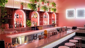 Una zona de bar con una barra larga, varios taburetes y estantes con botellas y plantas.