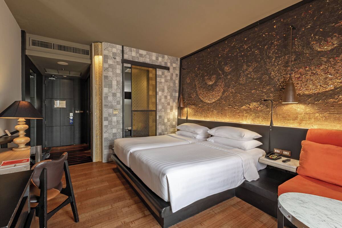 A hotel room with a bed, a desk, a chair, a nightstand, and a wall with a textured artwork.
