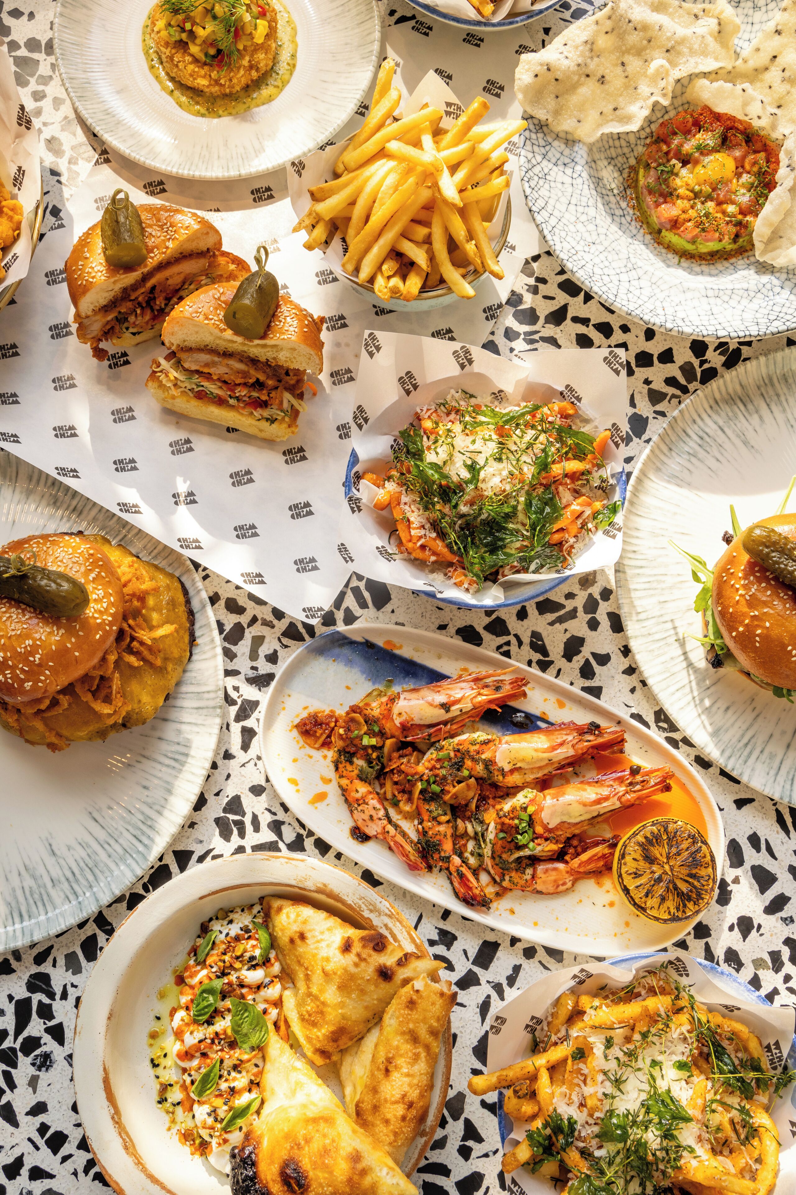 Une table où l’on trouve différents plats, notamment des hamburgers, des frites, des crevettes et des pâtes.