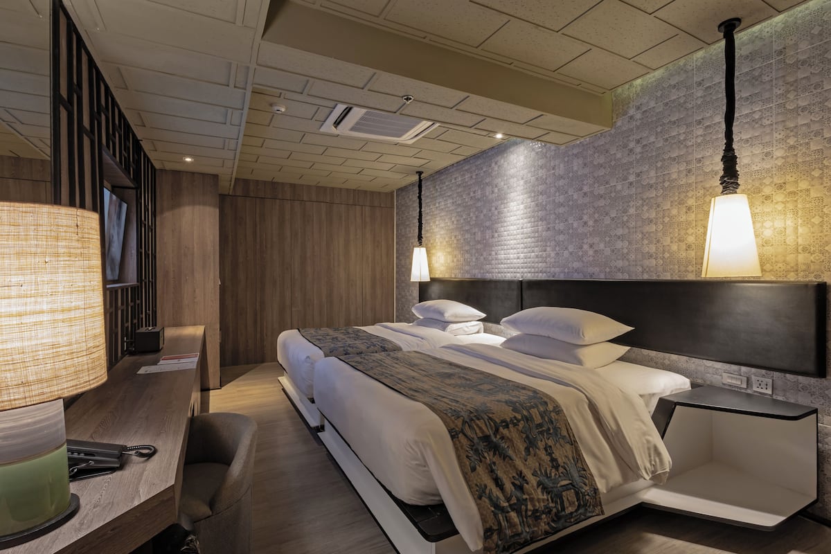 A modern hotel room with a large bed, bedside tables, a desk with a lamp, and a patterned wall.
