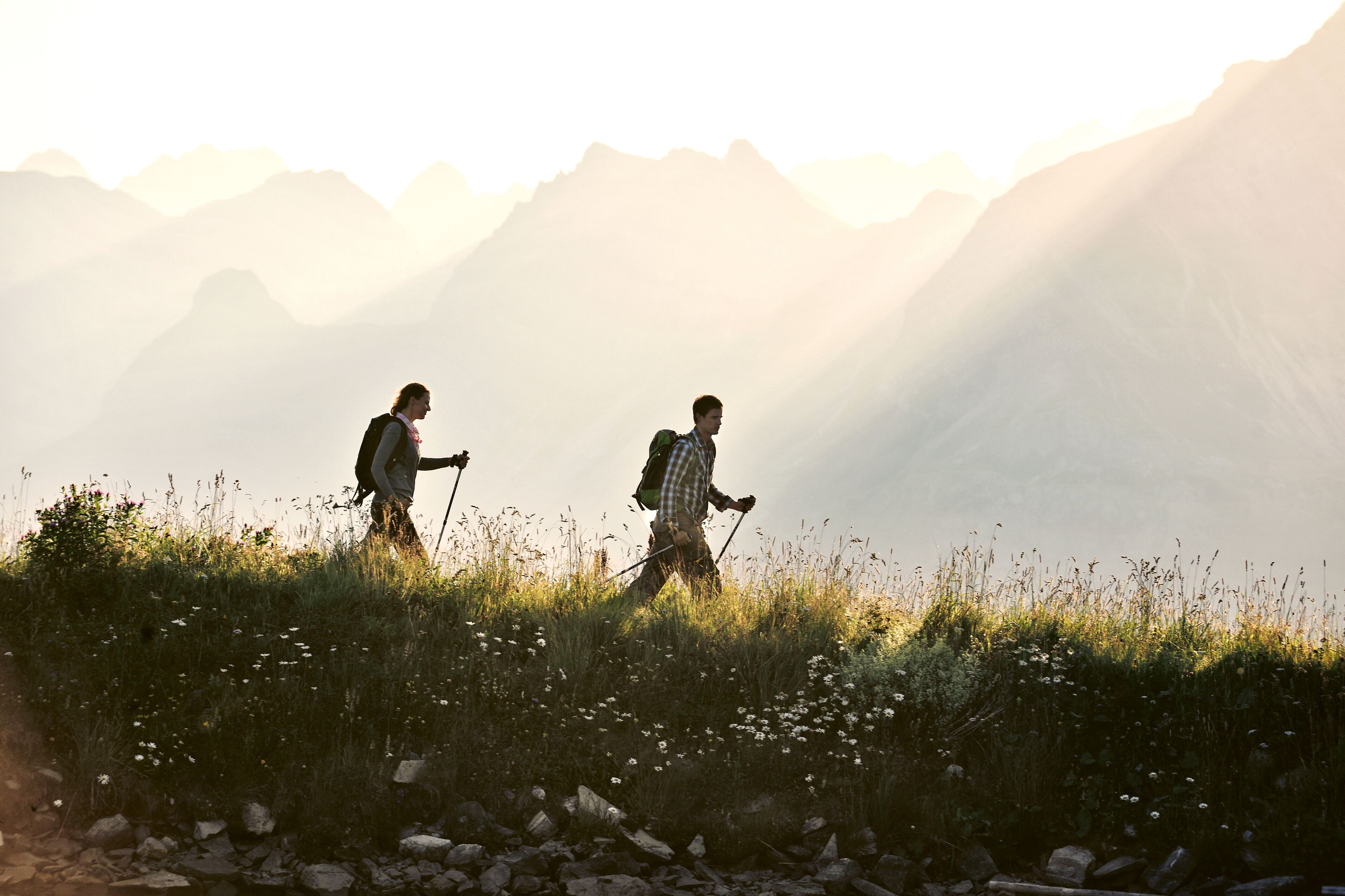 Twee wandelaars lopen over een begroeiing pad, met bergen op de achtergrond.