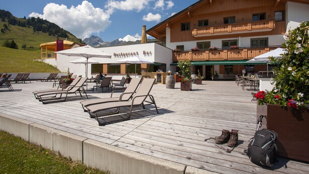 A wooden deck with lounge chairs and umbrellas, a restaurant bar, and a building with balconies. - Sporthotel Steffisalp (Warth)