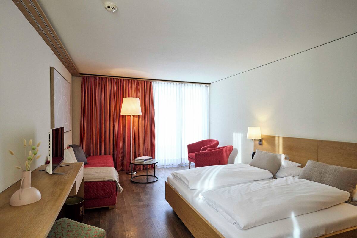 A hotel room with a large bed, a desk with a computer, a chair, a small table with a lamp, and a TV.