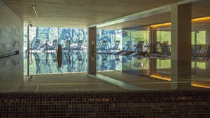 An indoor swimming pool with clear glass walls, multiple sun loungers, and a view of a forested area. - Sporthotel Steffisalp (Warth)