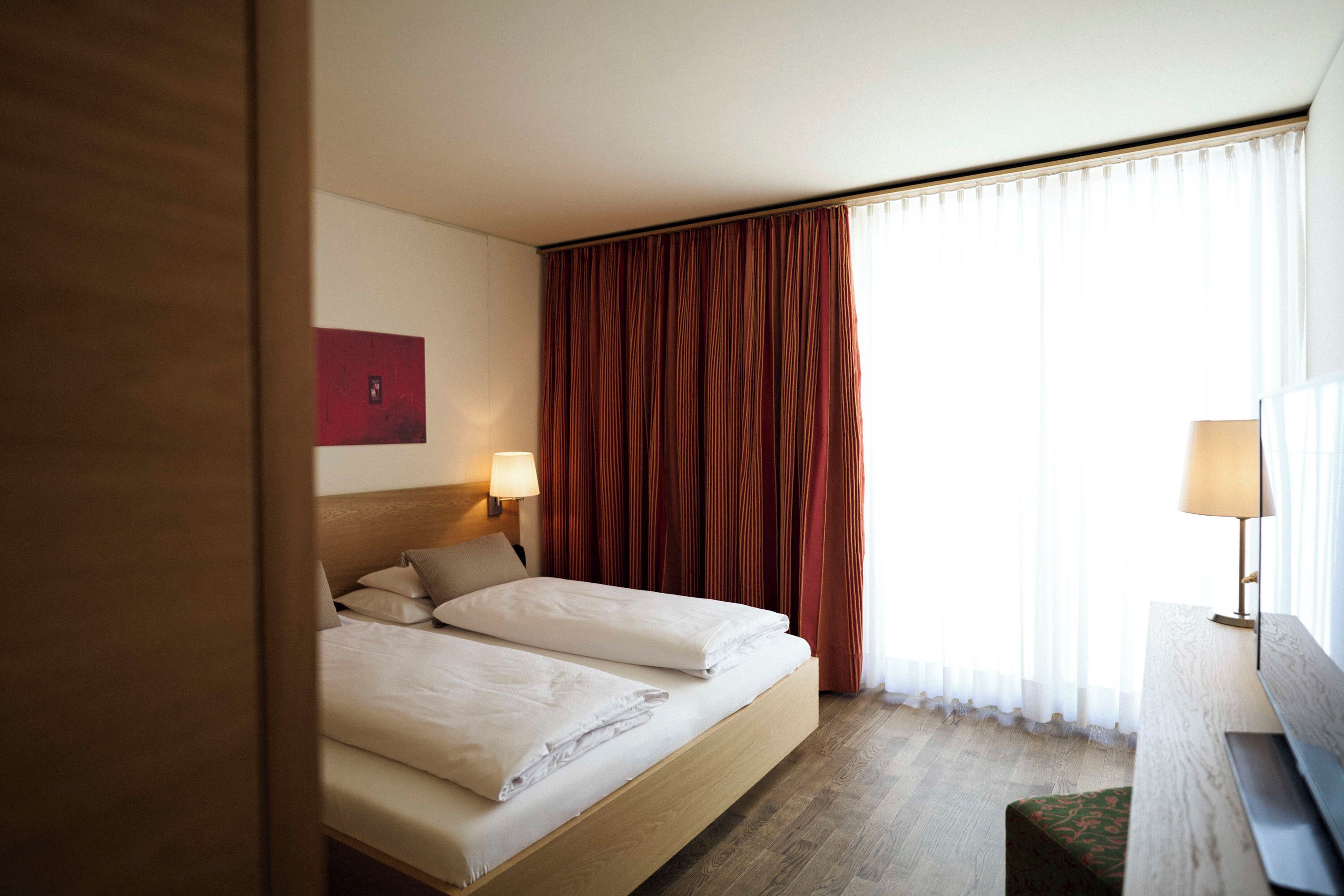 Ein Hotelzimmer mit zwei Betten, einem hölzernen Kopfteil, einem Nachttisch mit Lampe, einem großen Fenster mit Vorhängen und einer roten Akzentwand.