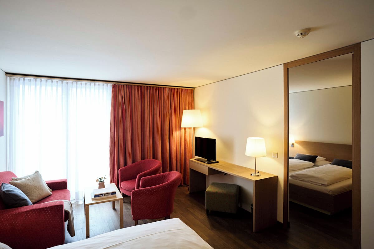 A hotel room with a bed, a desk with a computer, a chair, a small table with a book, and a TV.