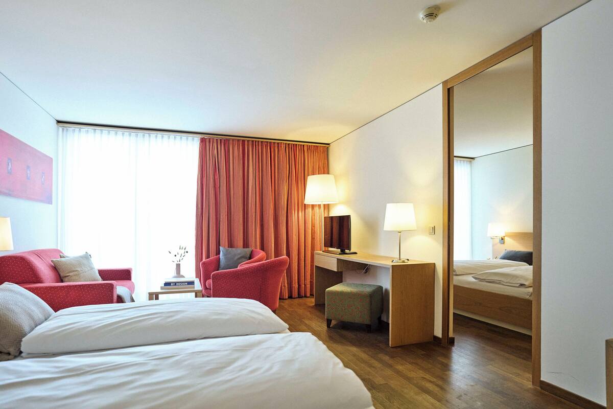 A hotel room with a bed, a desk, a chair, a small table with a lamp, and a large mirror.