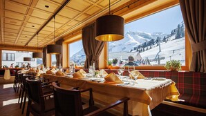 A restaurant with a long table set for dining, large windows with a mountain view, and wooden ceiling panels. - Sporthotel Steffisalp (Warth)