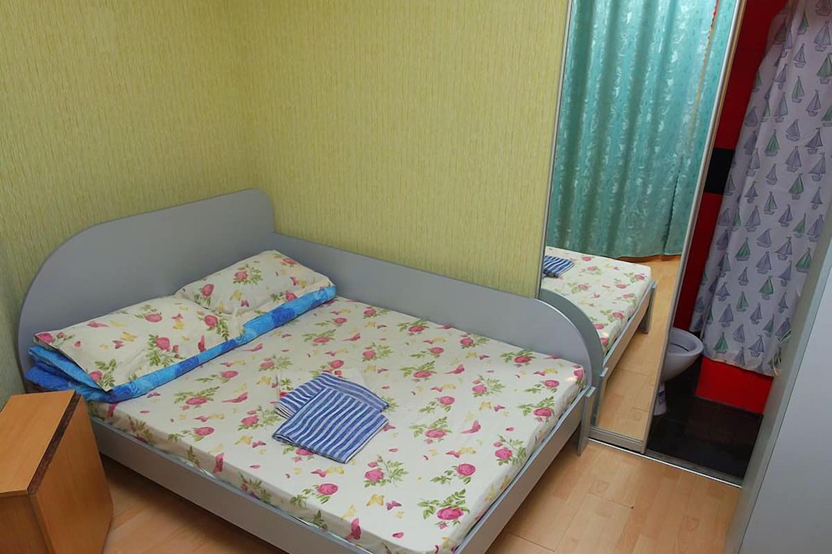 A small, single-bed room with a wooden floor, a grey bed frame, floral bedding, and a mirror on the wall.