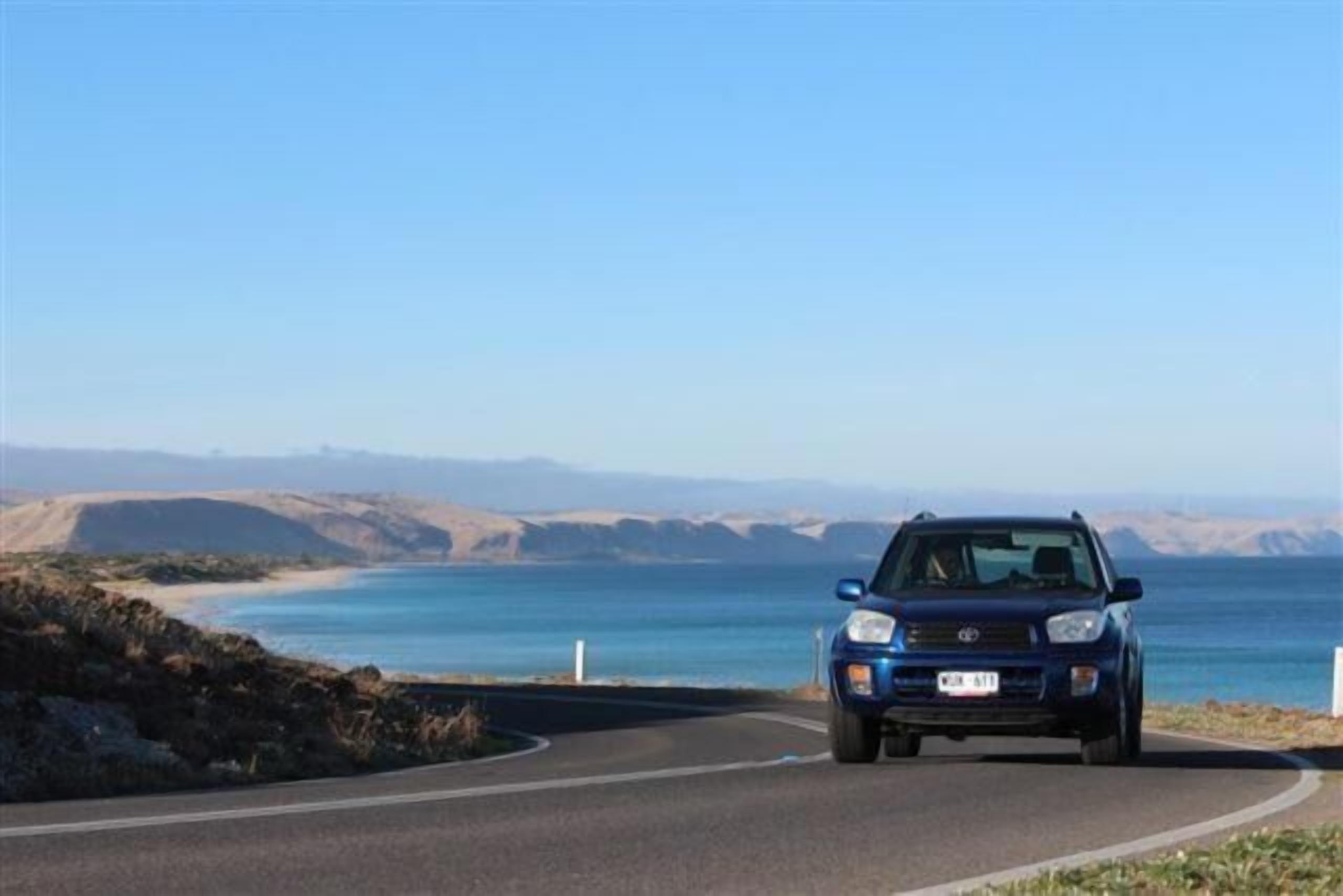A blue SUV driving on a coastal road with a clear view of the ocean and distant hills.