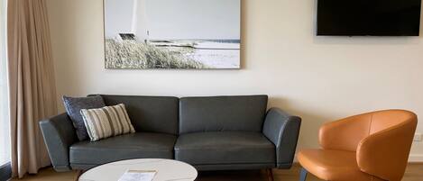 A living room with a grey sofa, an orange chair, a small white table, a framed picture of a lighthouse, and a flat-screen TV on the wall.
