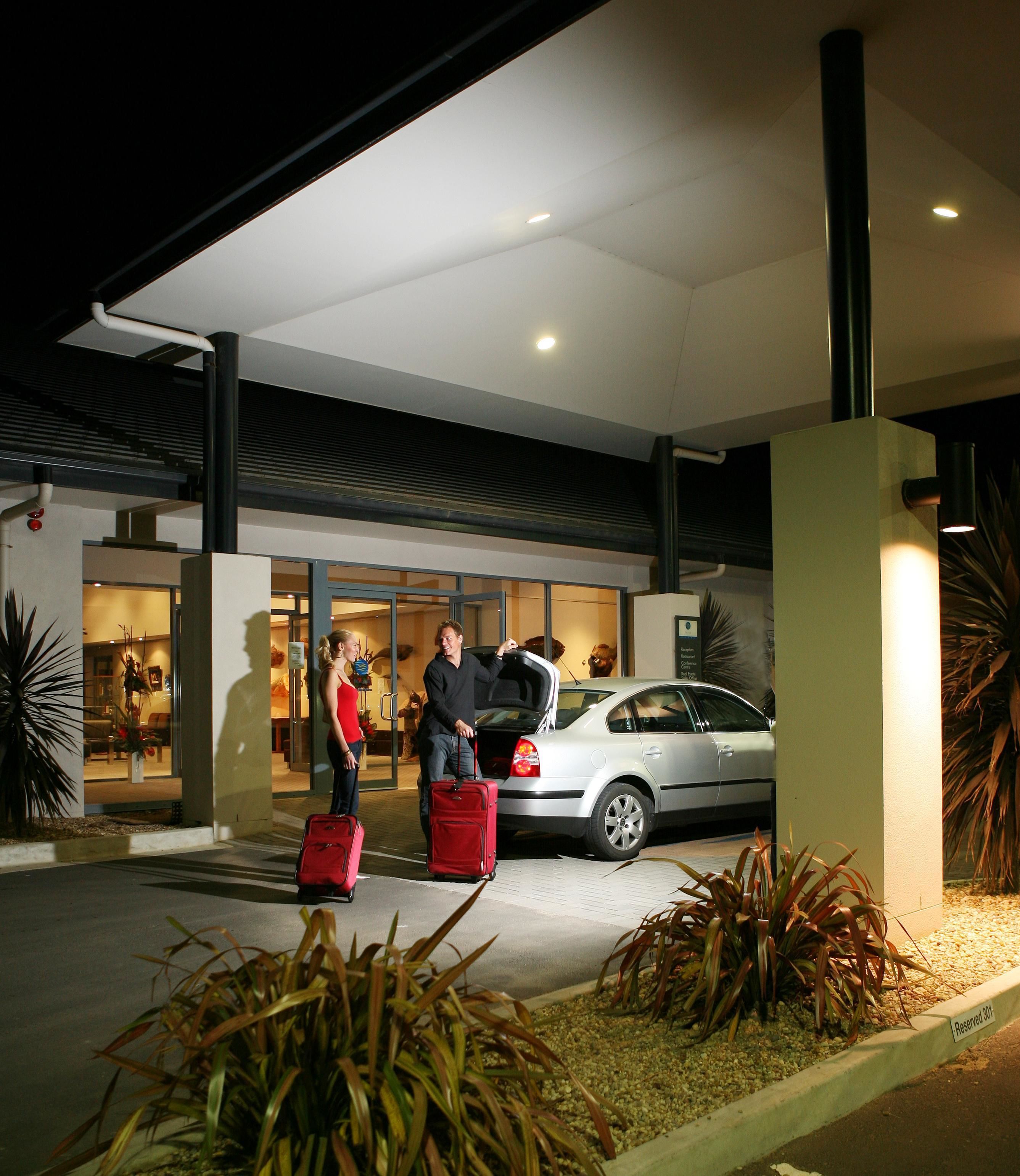 A modern building with a covered entrance, a car parked in front, and two people with luggage.