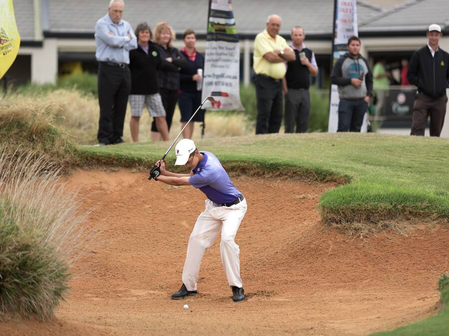 Un golfista con camisa púrpura y pantalones blancos a punto de golpear una pelota desde un búnker de arena.