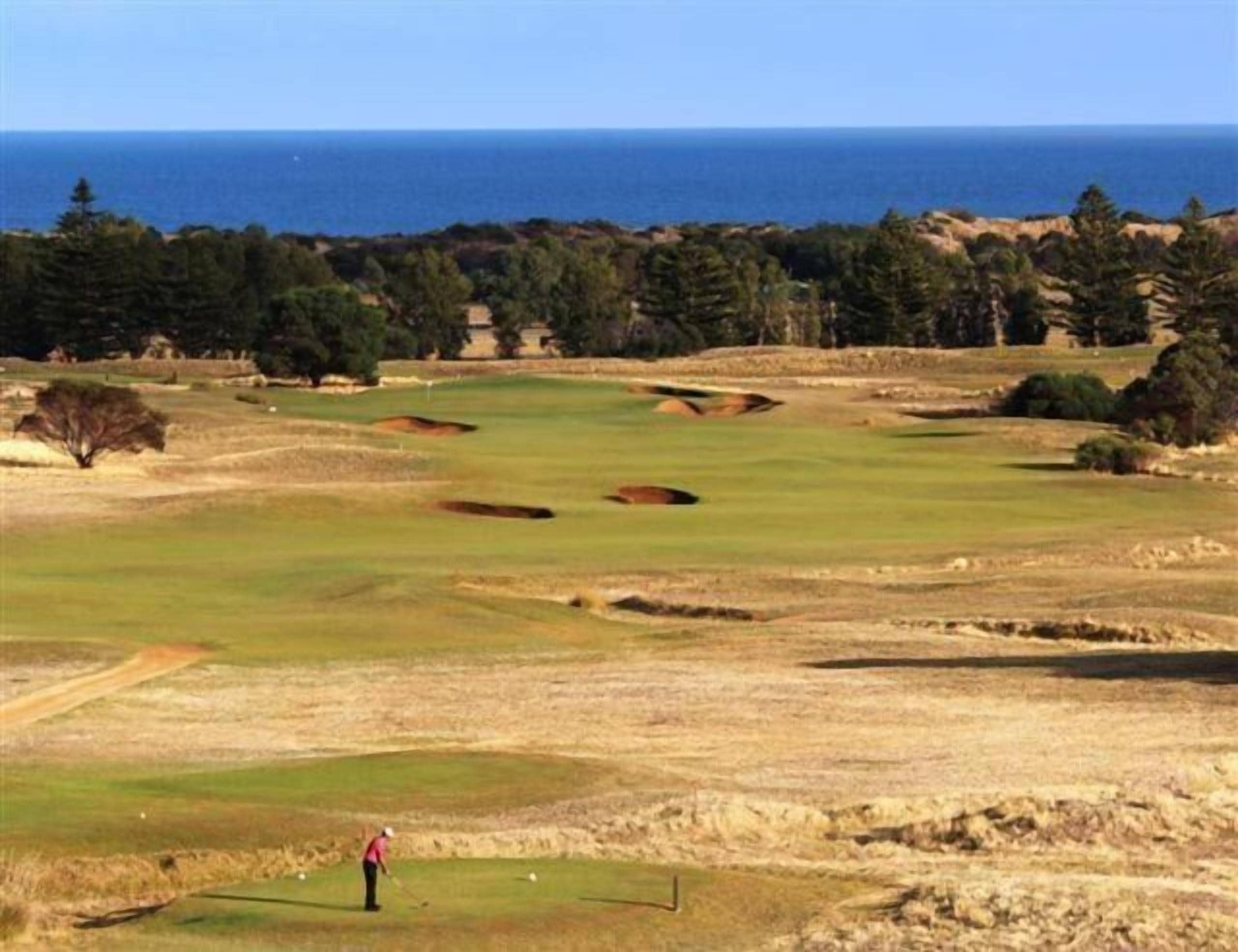 Um campo de golfe com um jogador, armadilhas de areia e vista para o oceano.