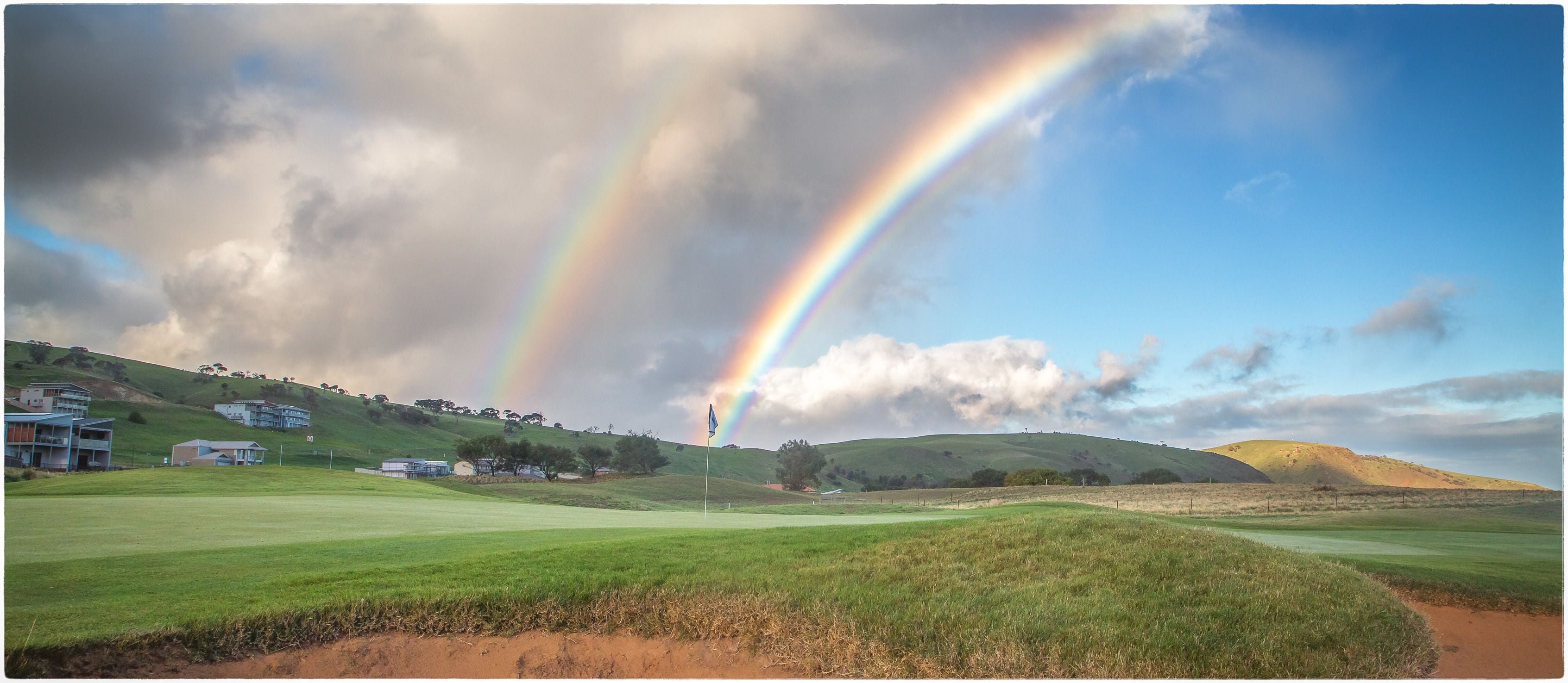 A golf course with a rainbow, houses on a hill, and a flag.