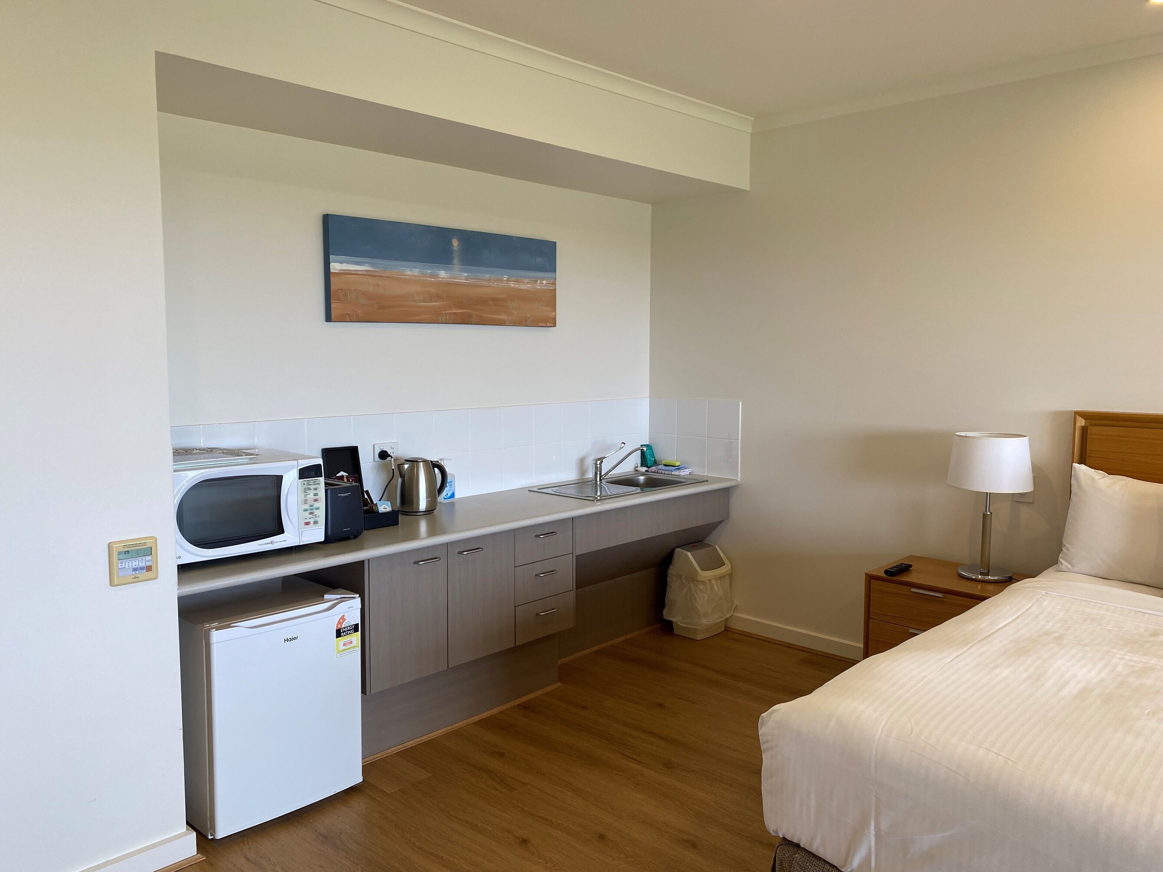 A compact hotel room with a kitchenette, a bed, a nightstand, and a painting on the wall.