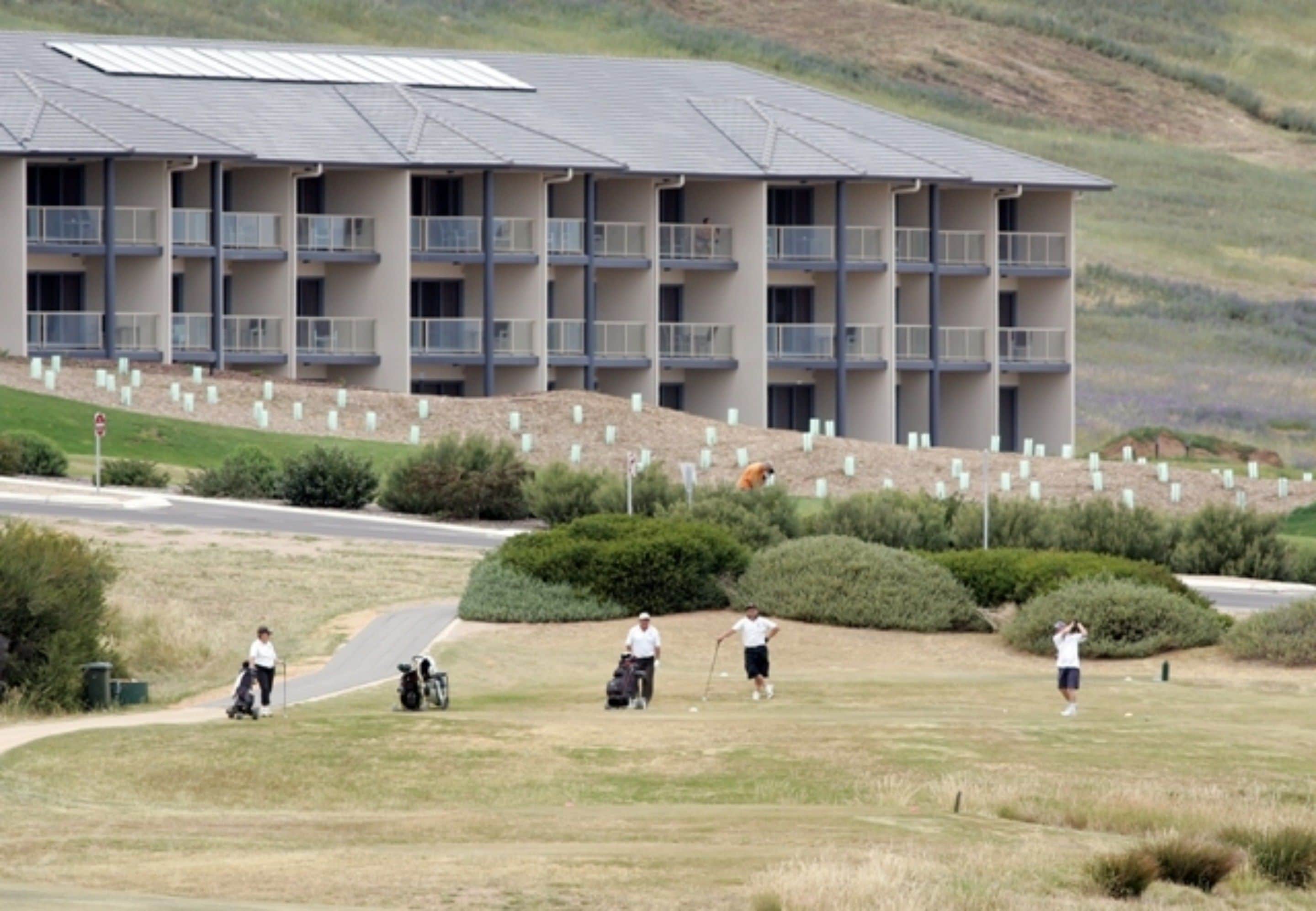 Un edificio de varios pisos con balcones, un campo de golf en primer plano y un paisaje montañoso al fondo.
