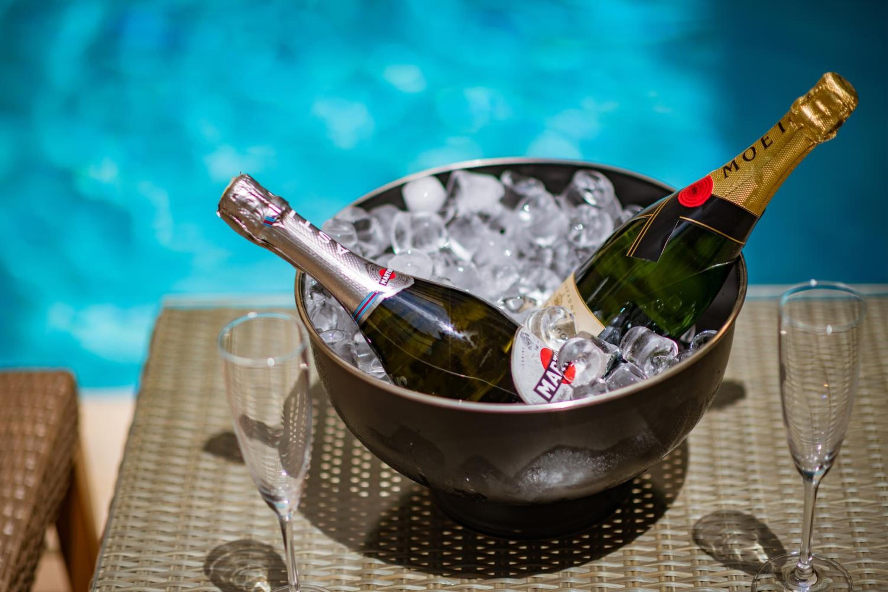 A bowl of ice with two champagne bottles and glasses on a table by a pool.