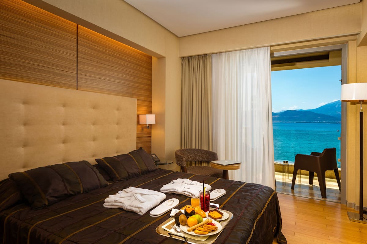 A hotel room with a large bed, a dining table with breakfast, a chair, and a view of the sea.
