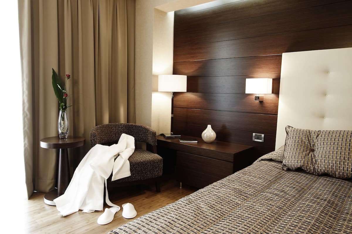 A hotel room with a bed, a chair, a nightstand, a lamp, and a vase with flowers.