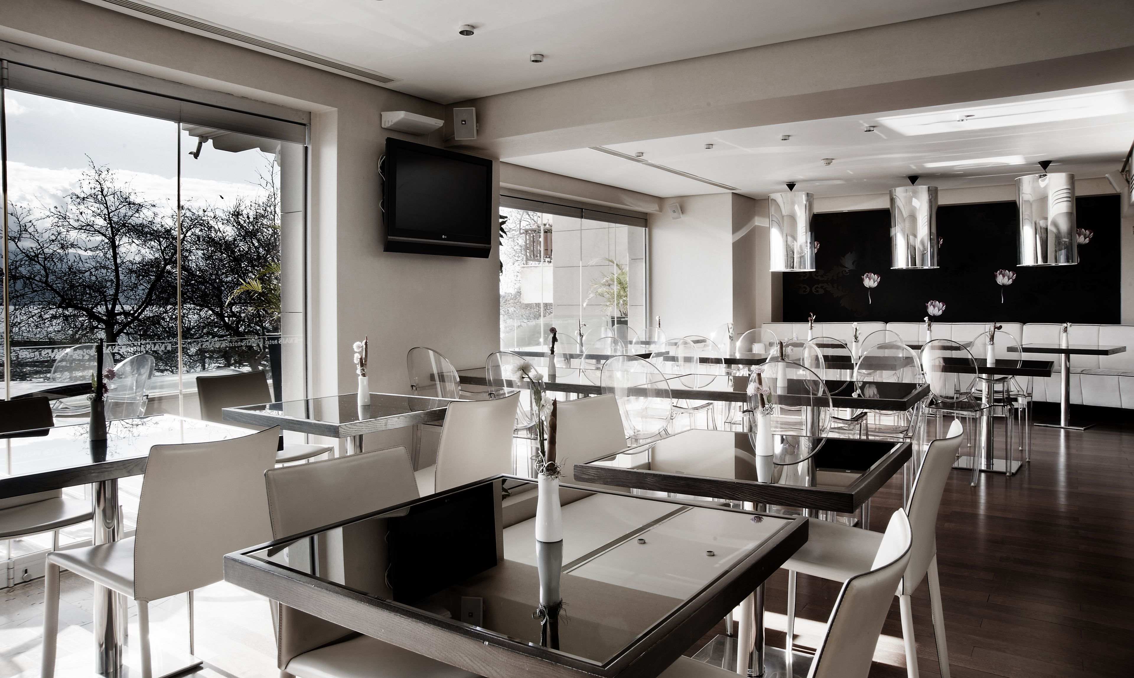 A modern dining area with glass tables and chairs, a black kitchen in the background, and a large window with a view of trees.