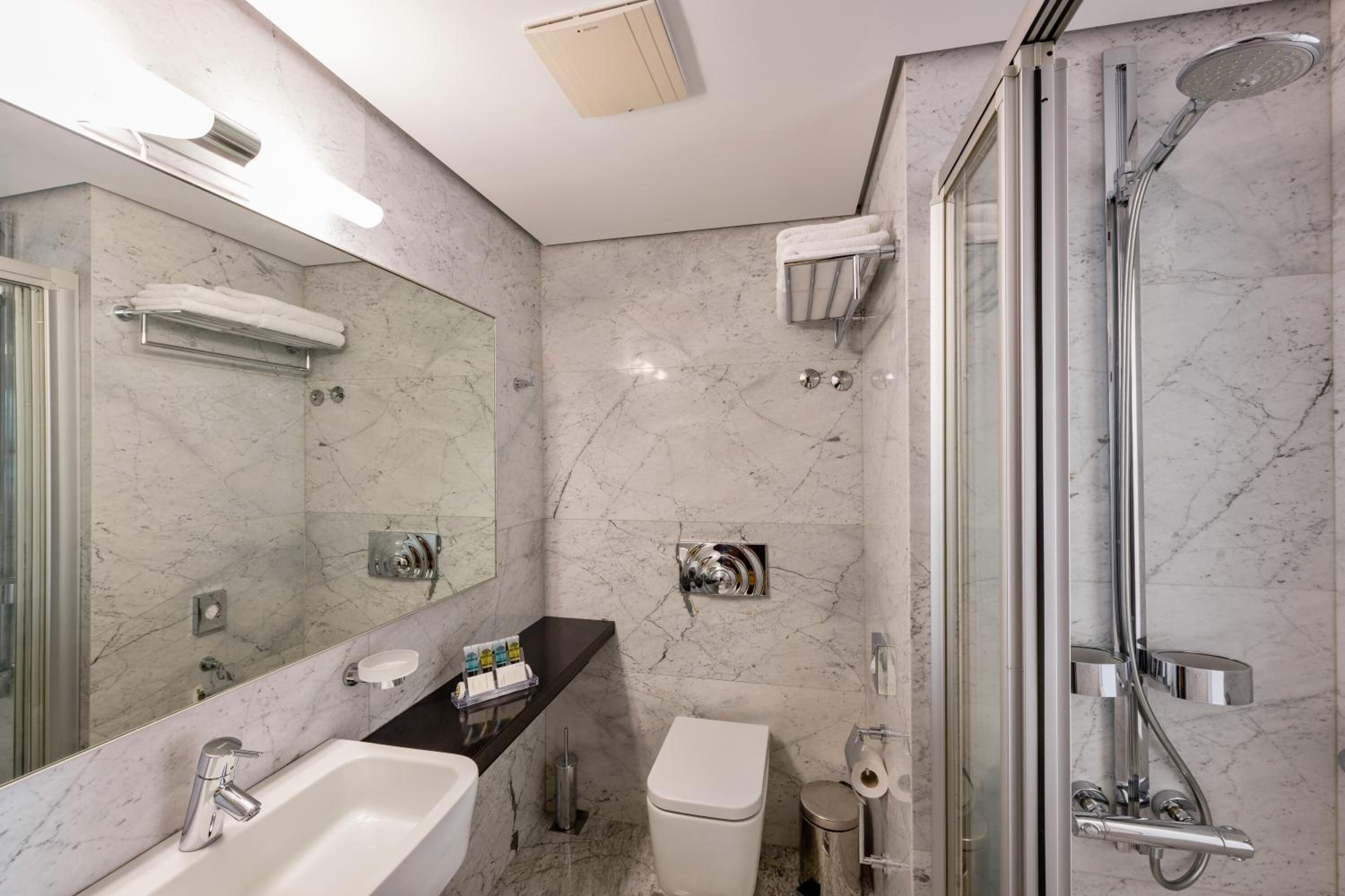A modern bathroom with a marble wall, a large mirror, a sink, a toilet, and a glass-enclosed shower area.