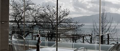 Un área de estar moderna al aire libre con mesas y sillas de vidrio transparente, con vista a un cuerpo de agua y árboles.