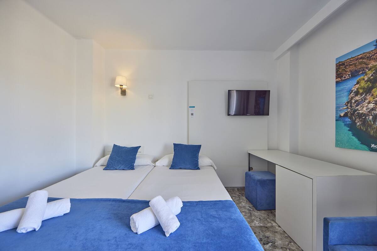 Une chambre d’hôtel avec deux lits, un bureau, une télévision et un tableau représentant un paysage côtier.