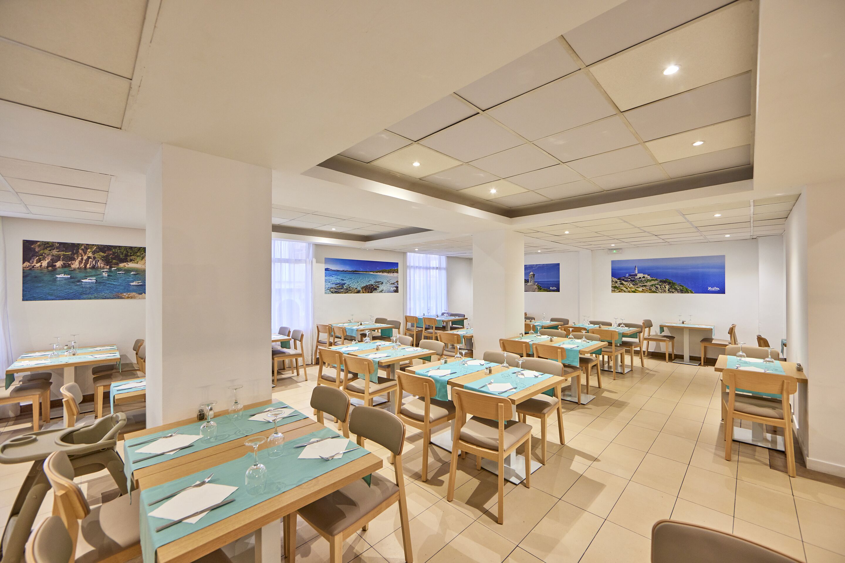 Spaziosa sala da pranzo con diversi tavoli e sedie, ampie finestre vista mare e opere d'arte raffiguranti scene costiere.