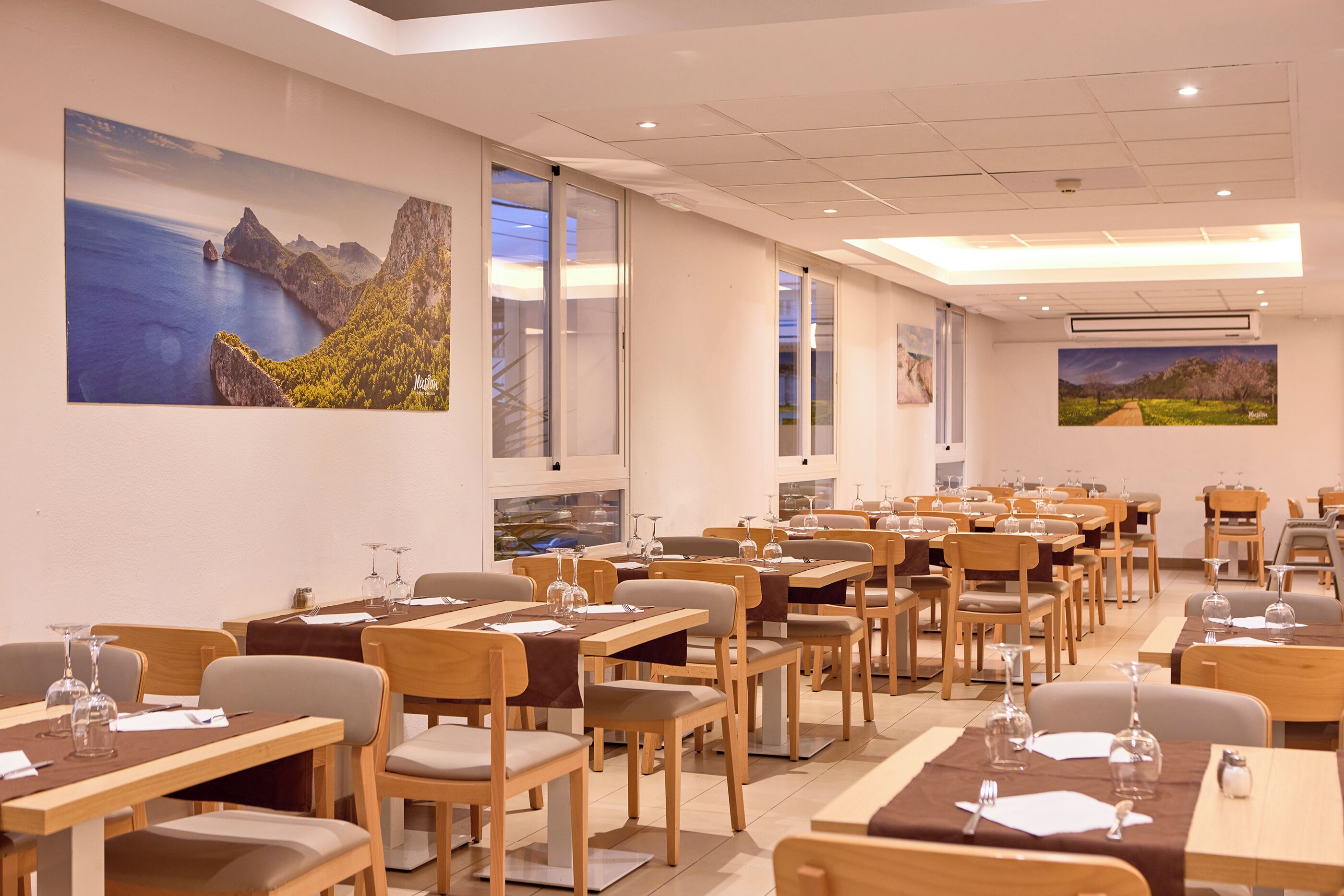Un restaurante con mesas y sillas de madera, ventanas amplias y obras de arte en las paredes.