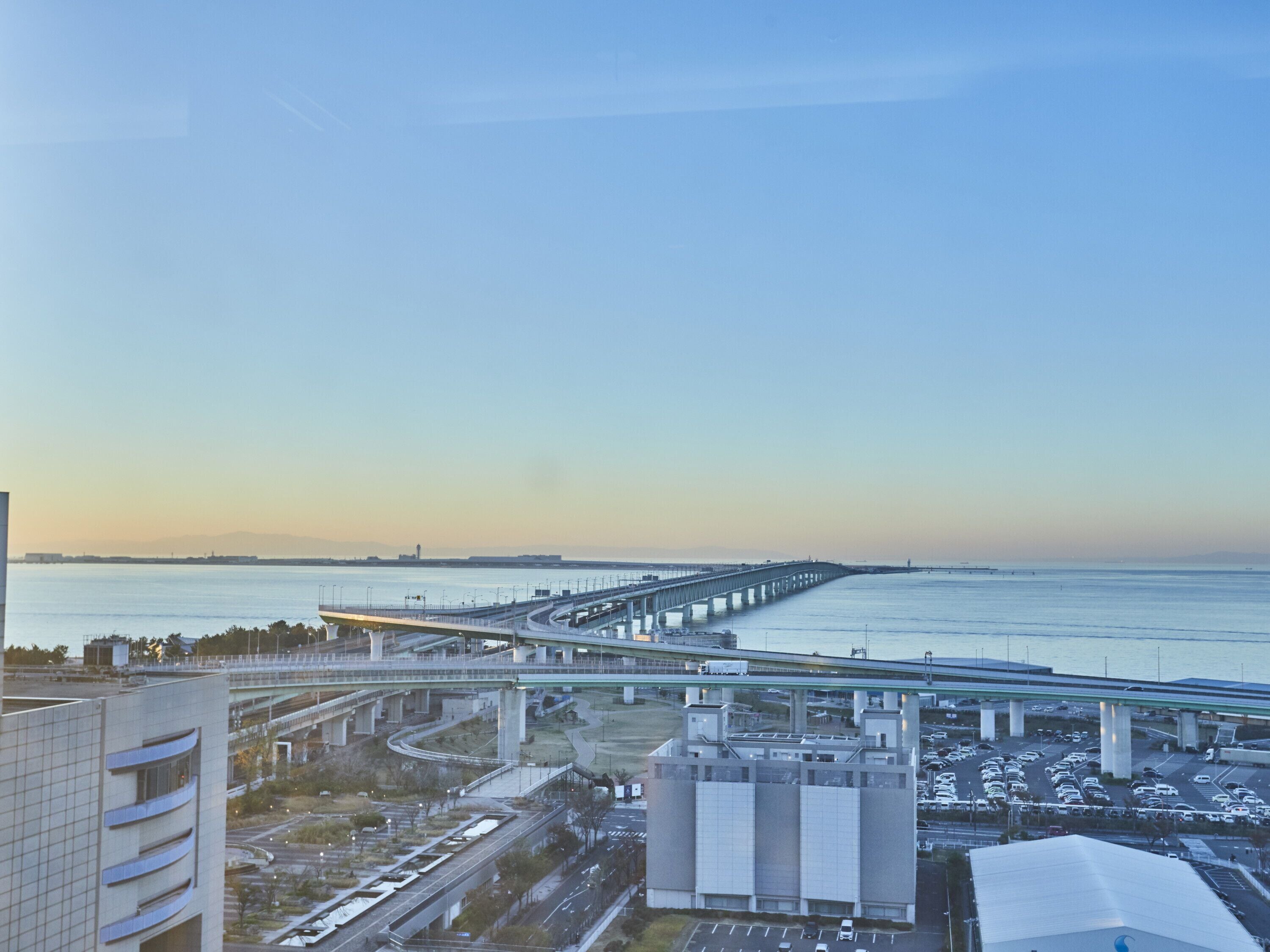 A coastal cityscape with a prominent bridge, modern buildings, and a parking area.