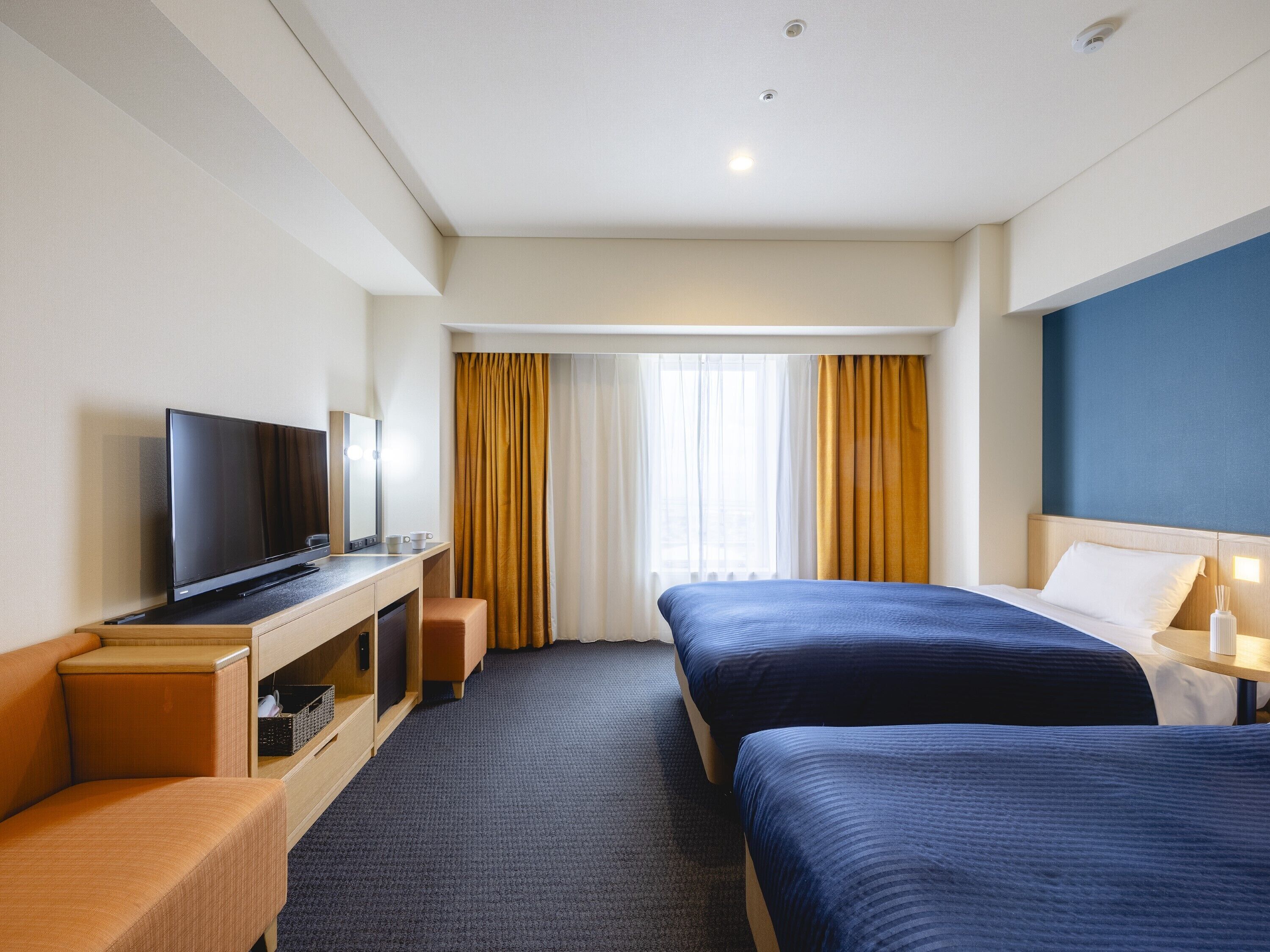 A hotel room with two beds, a TV, a desk, and a window with curtains.
