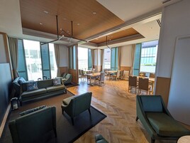 A spacious hotel lobby with a modern design, featuring a seating area with sofas and chairs, a dining area with tables and chairs, and large windows providing natural light.