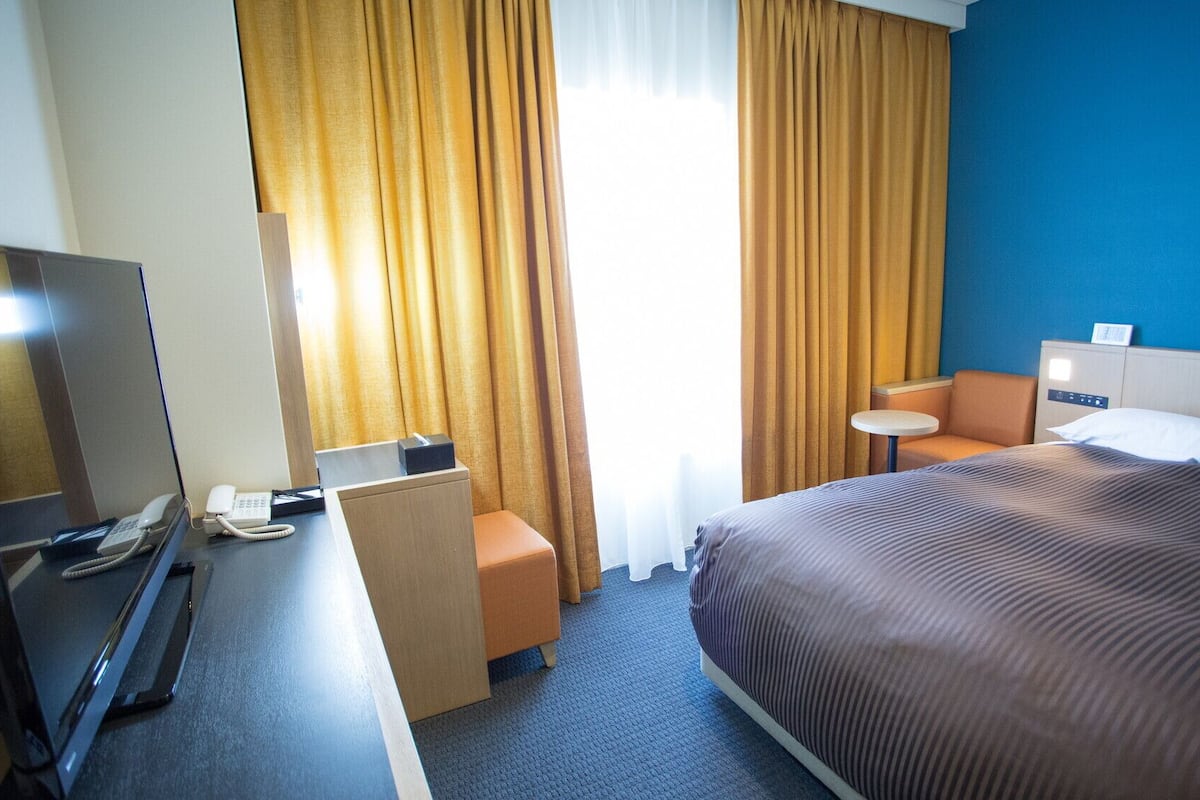 Ein Hotelzimmer mit einem Bett, einem Schreibtisch mit Telefon, einem Sessel, einem kleinen Tisch und einem Fernseher.