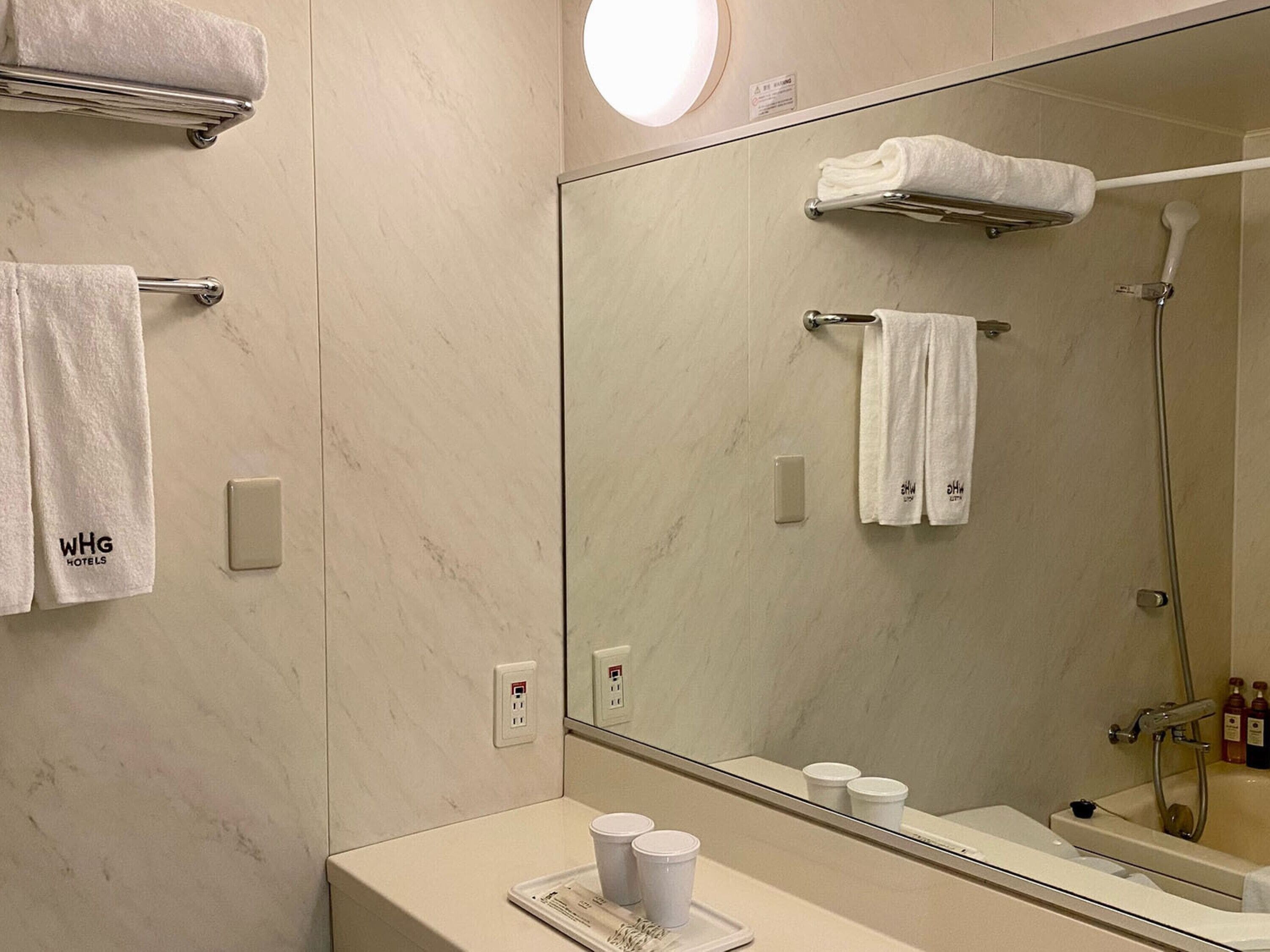 A hotel bathroom with a large mirror, two towel racks, a sink with toiletries, and a shower.