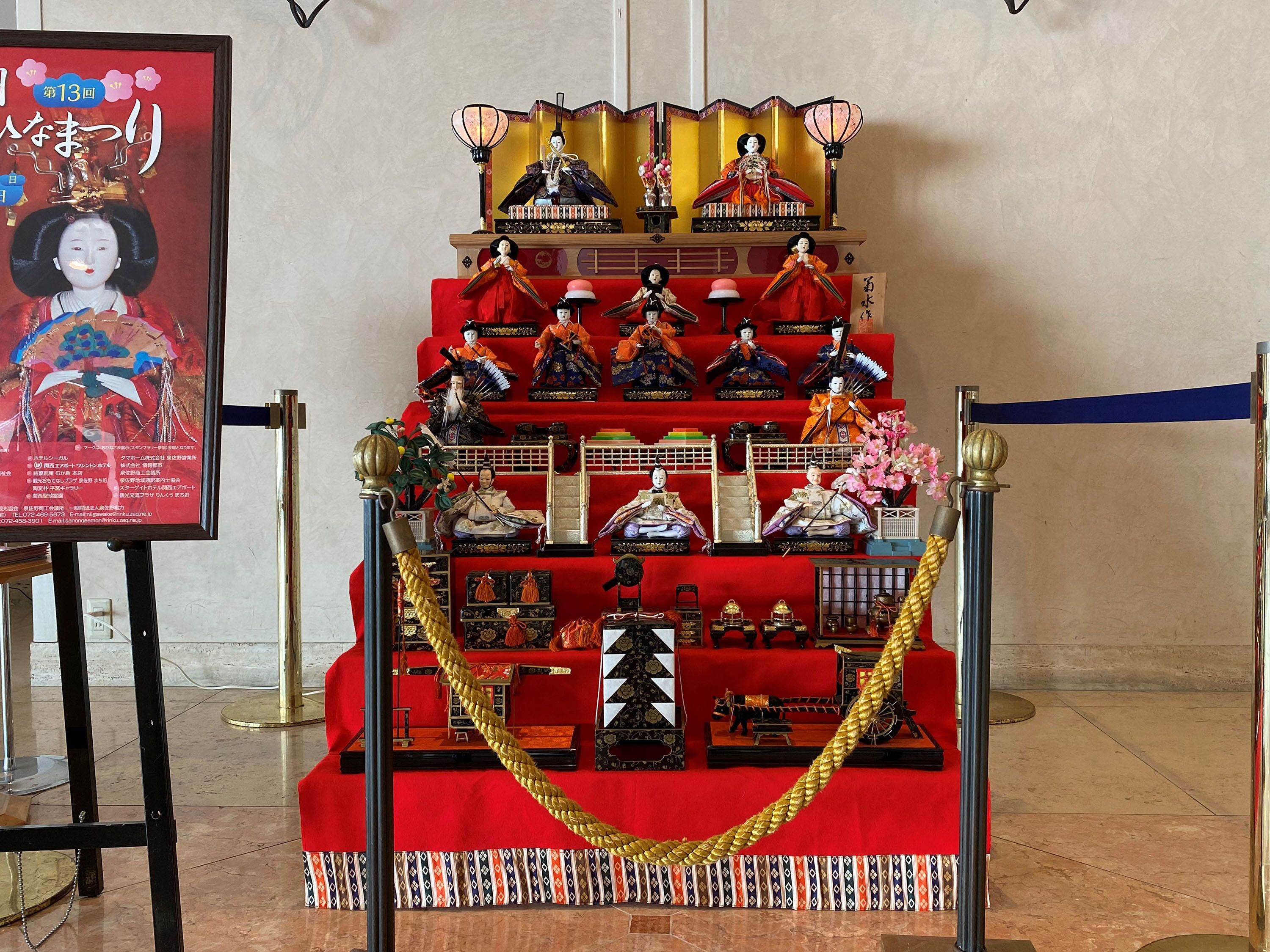 A display of traditional Japanese dolls and artifacts on a red carpet, with a golden rope barrier in front.