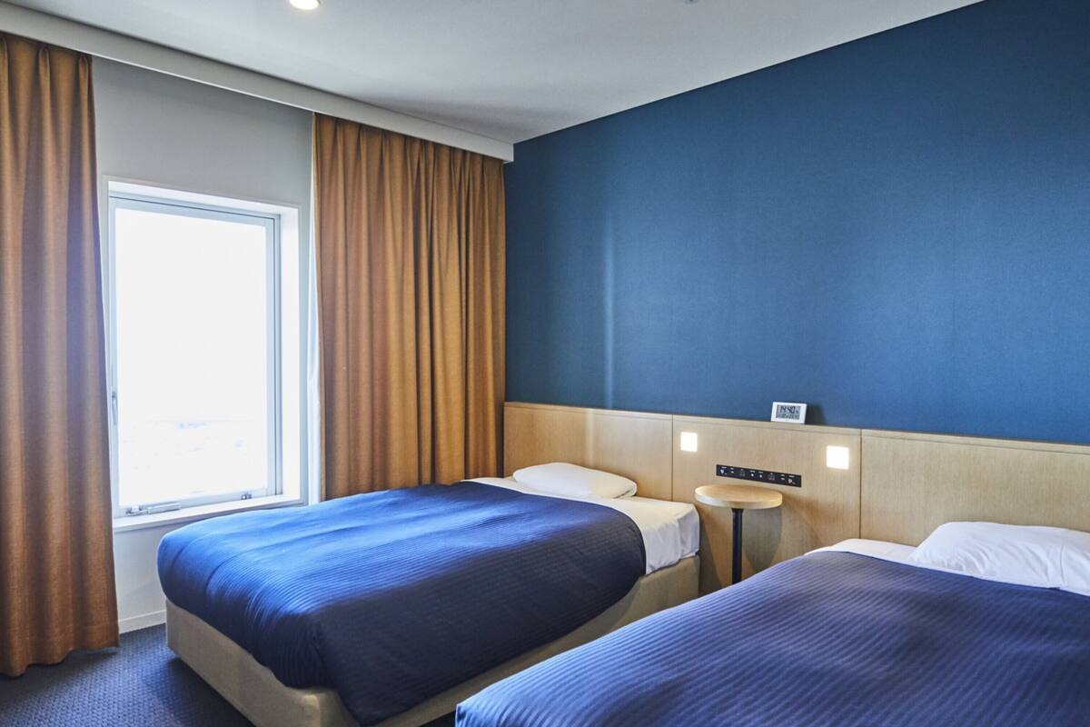 Een hotelkamer met twee bedden, een raam met gordijnen, een nachtkastje en een bedieningspaneel aan de muur.