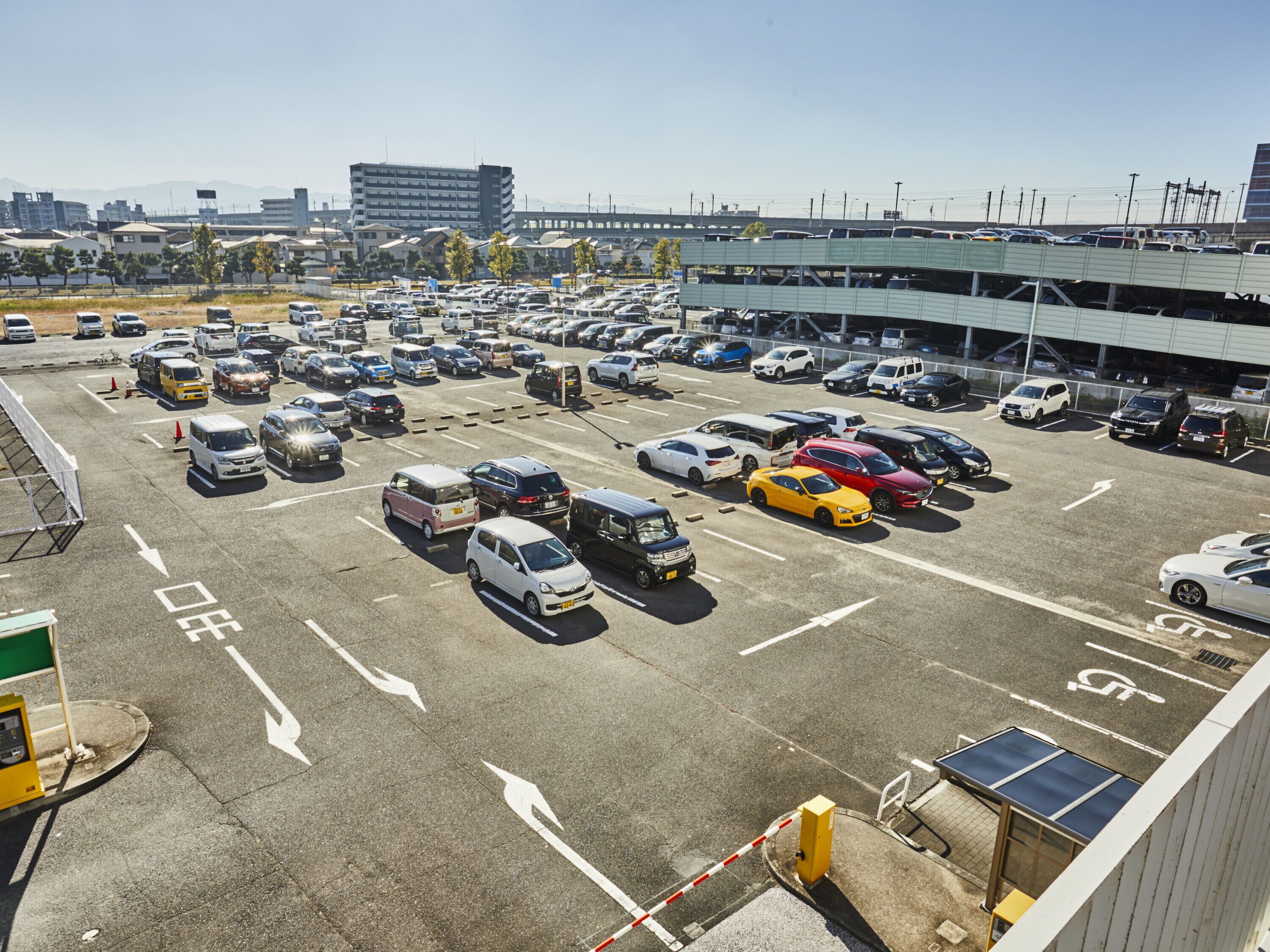 Un estacionamiento con varios vehículos, incluyendo automóviles, furgonetas y autobuses, y una estructura de estacionamiento de varios niveles al fondo.