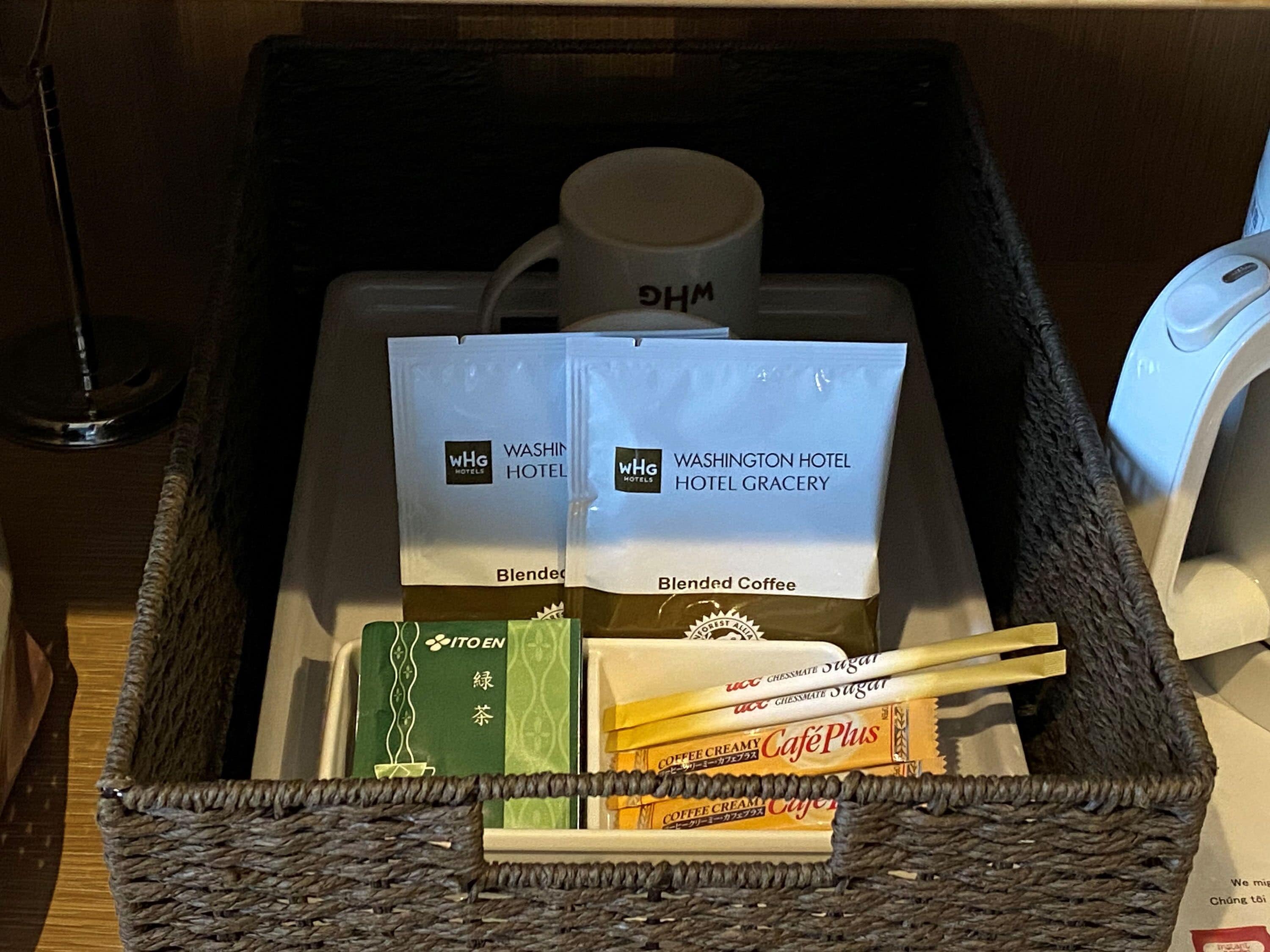 A hotel room's minibar with a coffee maker, two bags of blended coffee, a green tea box, and a box of Café Plus coffee cream.