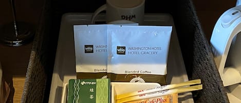 A hotel room's minibar with a coffee maker, two bags of blended coffee, a green tea box, and a box of Café Plus coffee cream.