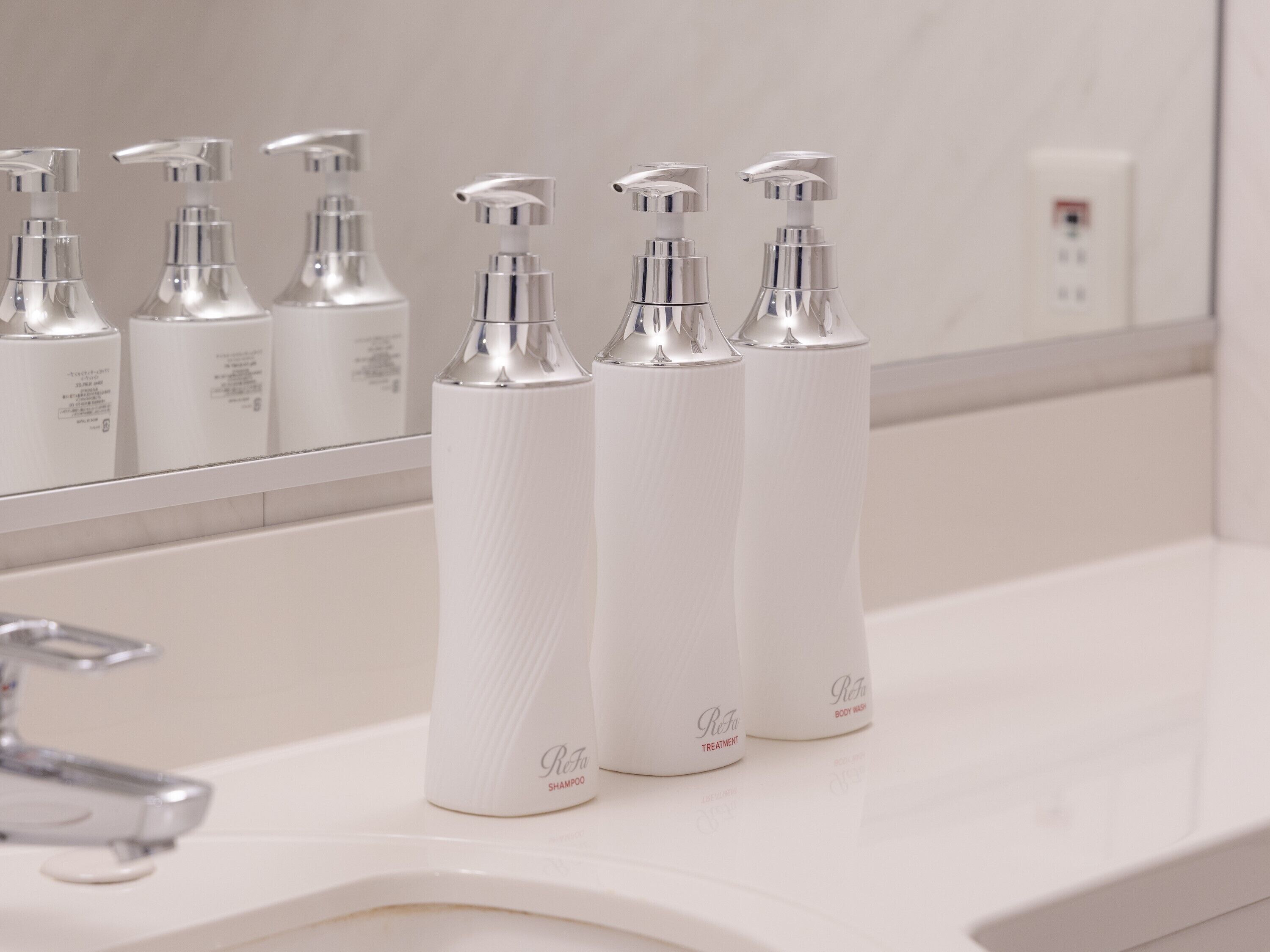 Une salle de bain avec un lavabo blanc, un miroir et trois flacons blancs de gel douche ReFa posés sur le comptoir.