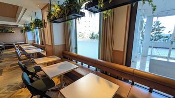 Un restaurant avec de longues tables en bois et des bancs rembourrés, de grandes fenêtres avec des rideaux et des plantes suspendues.