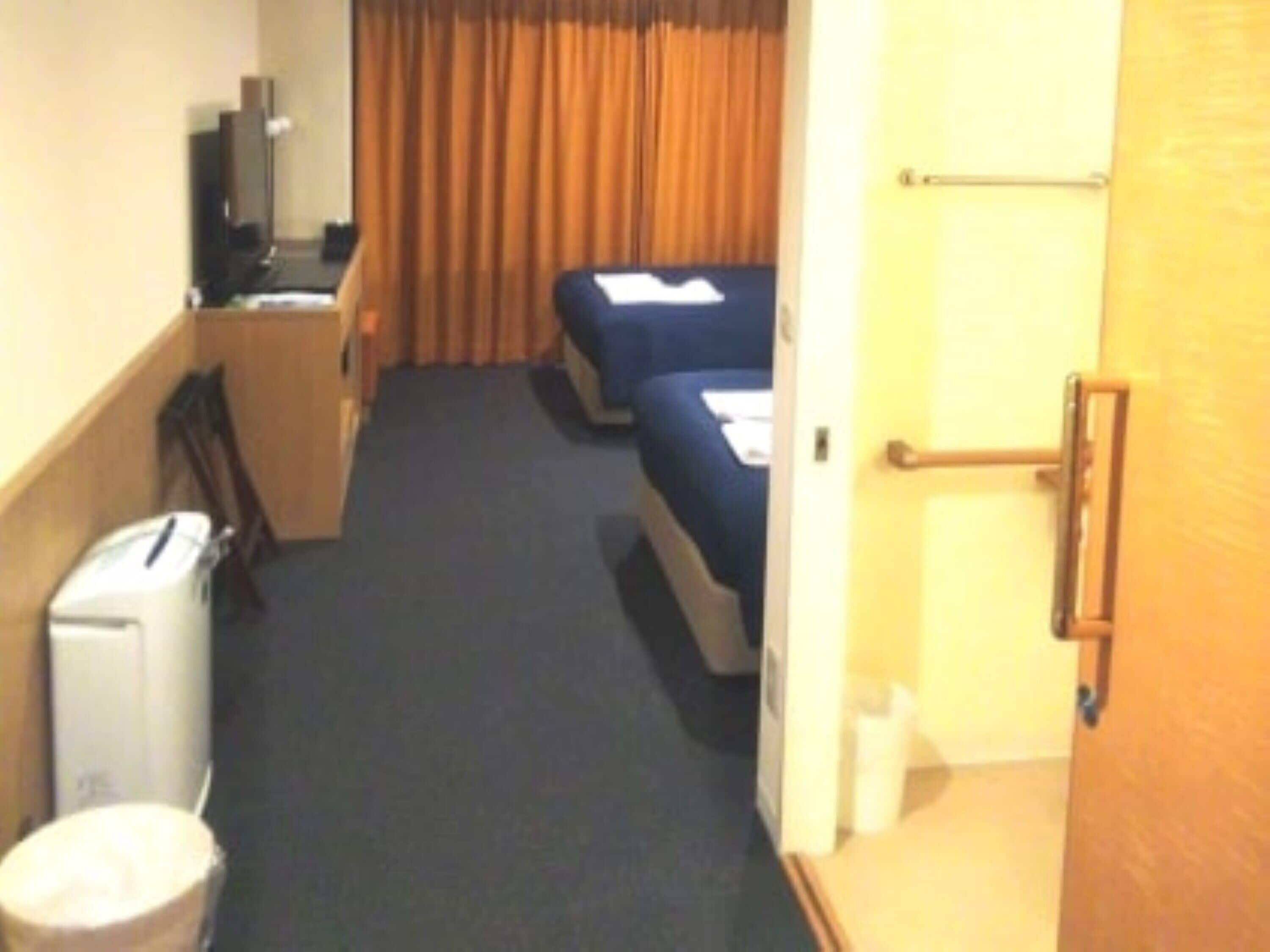 A narrow hotel corridor with a bed, a desk, and a trash bin.